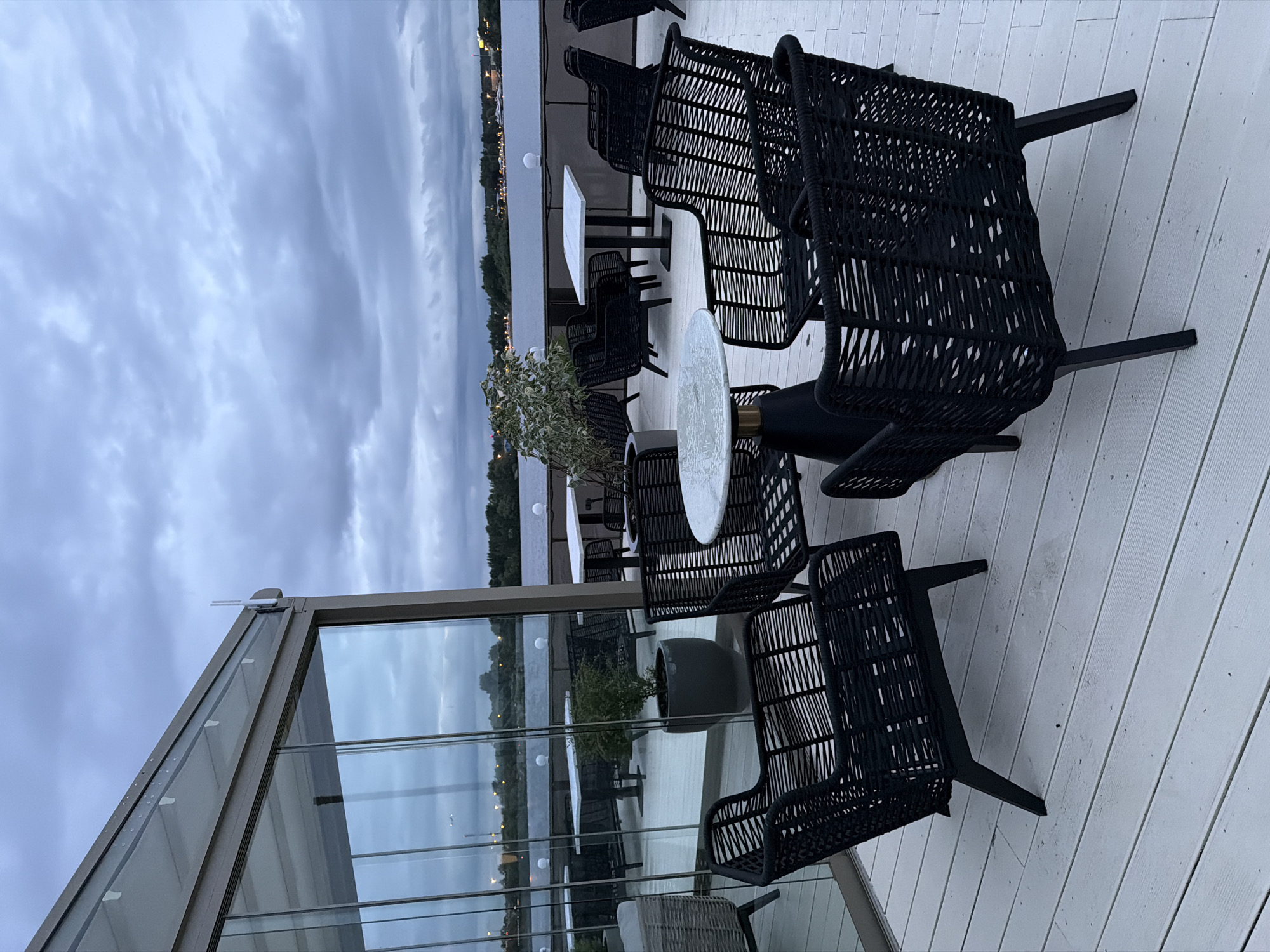 Captured at an indeterminate time of day in an unidentified city, the image presents an outdoor dining area. The scene is set on a wooden deck, where several black woven chairs are arranged around small tables. Adjacent to this area is a glass-enclosed structure, likely a building or conservatory. The reflections on the glass panes mirror the cityscape and the overcast sky. Beyond the deck, the cityscape is visible, marked by lights and structures along the horizon. The dominant element is the sky, heavy with clouds, suggesting an overcast or stormy weather condition. No individuals are visible in the frame.