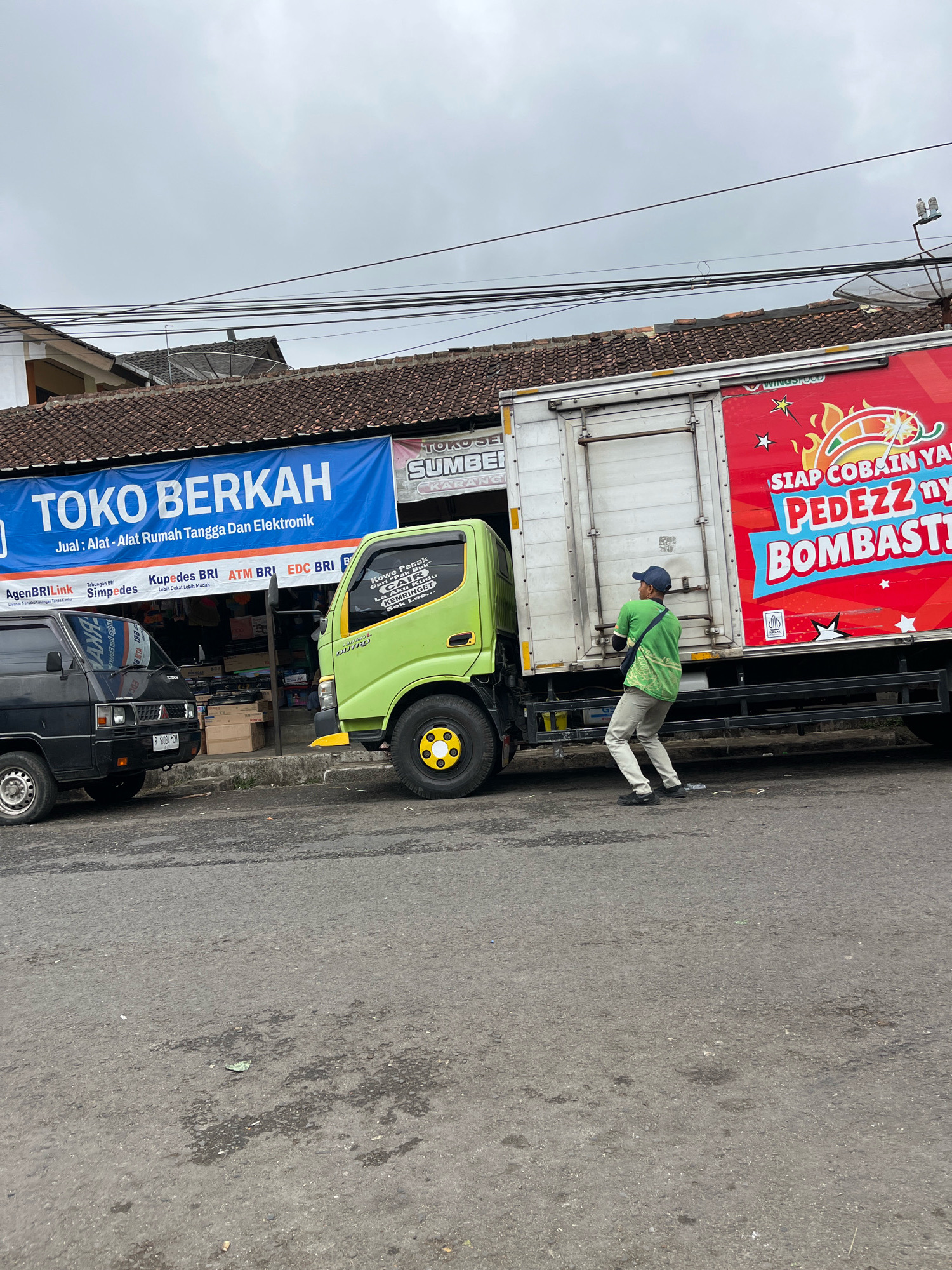 A man wearing a green t-shirt and a blue cap is pushing a light green truck. The truck is parked next to a black van, and both vehicles are in front of a storefront with a blue banner that reads "TOKO BERKAH" (meaning "Blessings Store"). The banner also states "Jual - Alat Rumah Tangga Dan Elektronik" (meaning "Sell - Household Appliances and Electronics"). The truck has a large red advertisement on its side featuring text in Indonesian, including "SIAP COBAIN YA PEDESZZ nya BOMBASTII". The scene appears to be on a street in Karangkobar, Indonesia, under an overcast sky. Wires are visible overhead.