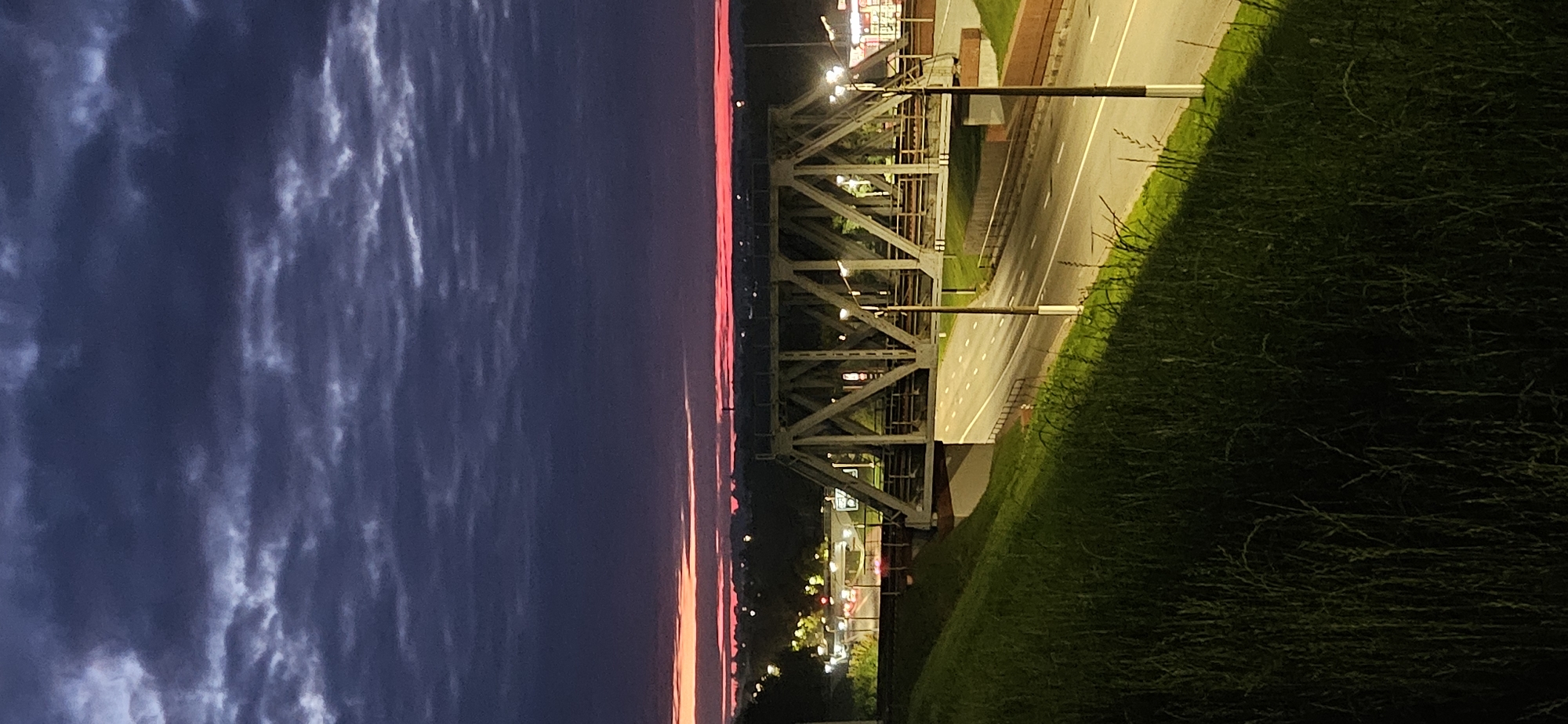 The image depicts a view of the railway bridge over the Vitebsk-Polotsk highway in Vitebsk, Belarus, at dusk. The sky is a deep purple with streaks of pink and orange near the horizon, indicating the setting sun. Dark, textured clouds fill the upper portion of the frame.
In the foreground, a steep grassy embankment slopes down from the left. The grass is tall and appears somewhat dark due to the low light conditions, with some areas illuminated by artificial light. A paved road runs horizontally across the mid-ground, with white lane markings visible. This is likely the Vitebsk-Polotsk highway.
Dominating the mid-ground is the prominent structure of a railway bridge. It is a steel truss bridge with a light grey or silver color. The bridge has a complex geometric design with numerous diagonal supports. Lights are visible along the bridge and on the road beneath it. Streetlights are also present along the road, casting a warm yellow glow.
In the background, a line of trees and buildings can be seen, with more lights suggesting an urban or suburban area. The overall atmosphere is calm and serene, characterized by the transition from day to night. No people are visible in the image. The scene is static and captures a specific moment in time at this location.