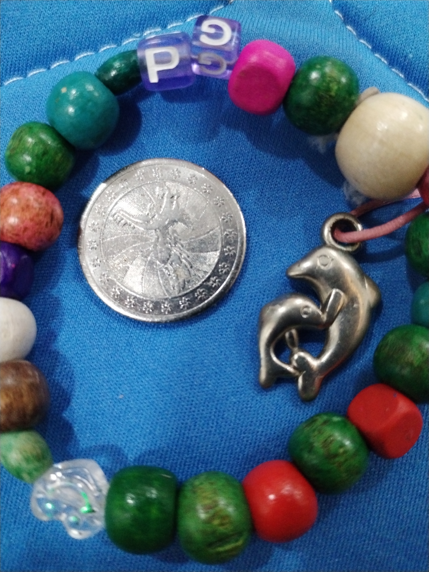 A close-up shot depicts a colorful bracelet on a blue fabric background, likely a piece of clothing. The bracelet is composed of various wooden beads in shades of green, brown, red, pink, white, and blue. Several lettered beads spell out "PS" on the upper section of the bracelet. A silver-colored coin, possibly a Euro, rests within the circle formed by the bracelet. A dolphin charm dangles from the bracelet, featuring two dolphins intertwined. The fabric beneath has subtle stitching detail. The photograph suggests a personal or sentimental item. While no specific location within Nha Trang Ward, Vietnam, is discernable, the shot appears to be taken indoors, possibly in a home.