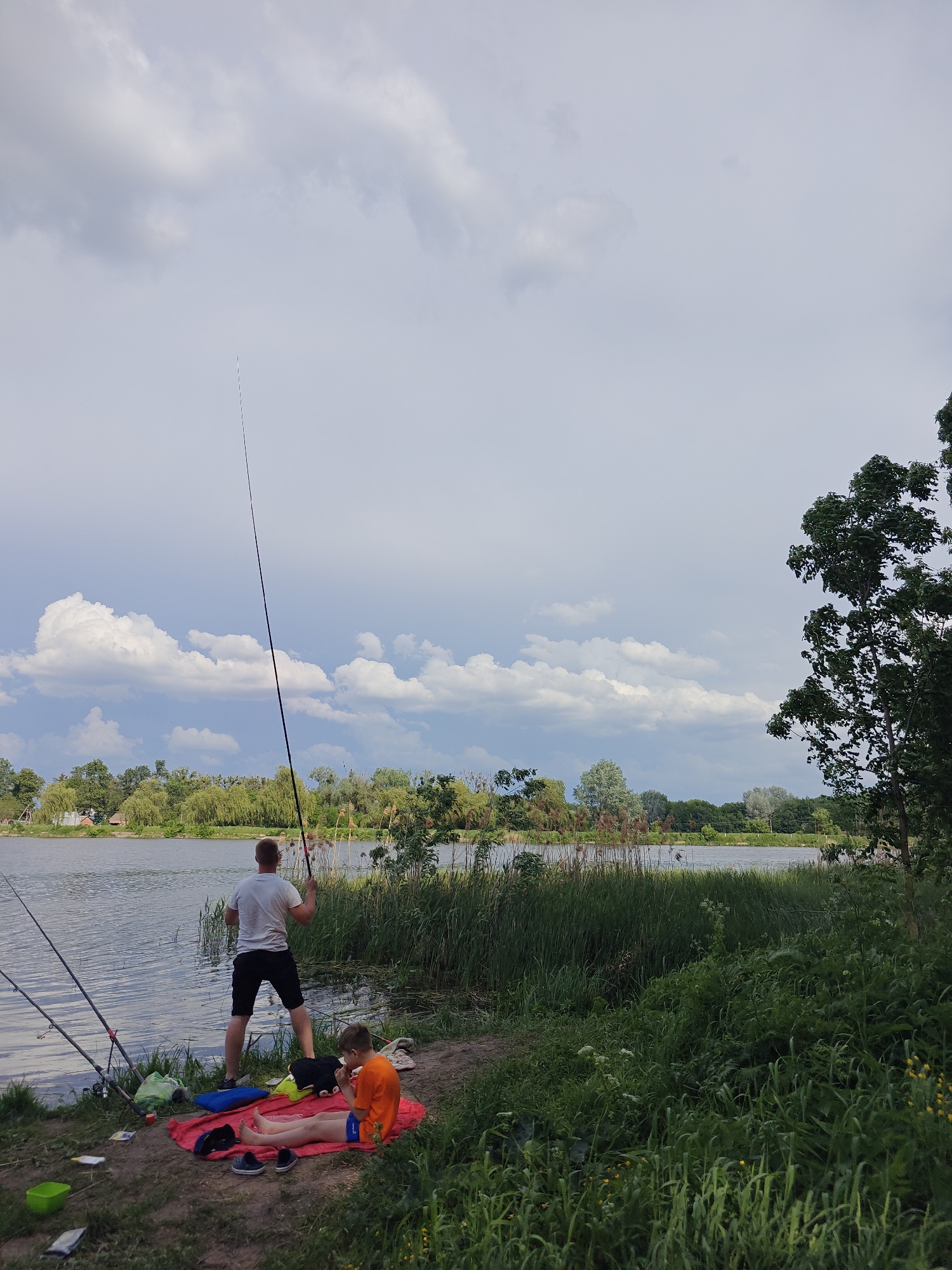 The image depicts a scene at a lake in Bobrivnyk, Ukraine. A man stands near the water's edge holding a fishing rod, while another fishing rod is propped nearby. A boy is seated on a red blanket, appearing to eat something. There are other objects scattered around the blanket, including shoes and a green container. The background shows the lake, trees, and a cloudy sky. Vegetation grows along the lake shore.
