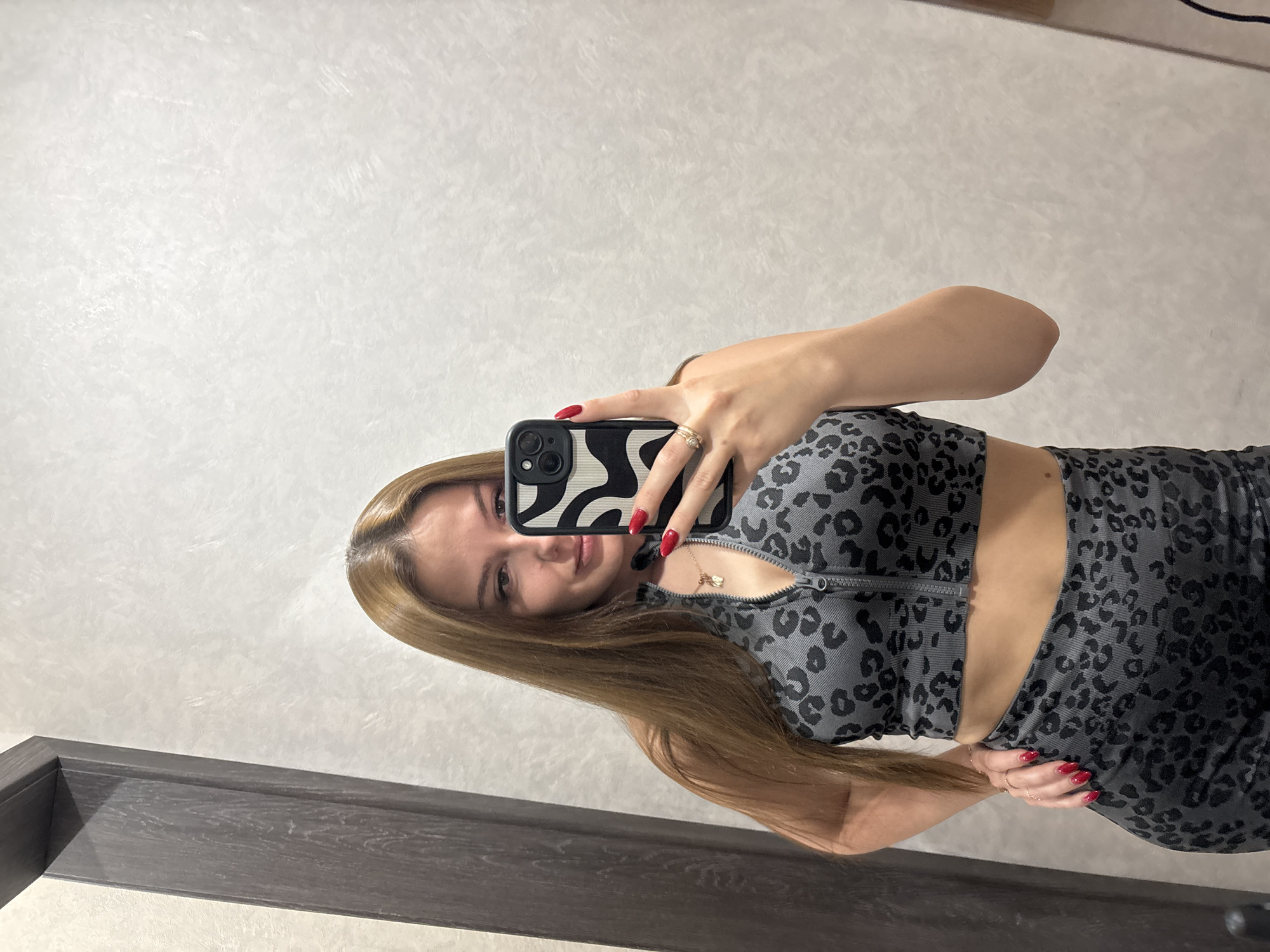 In a dimly lit indoor setting, a woman with long, light brown hair is taking a selfie in a mirror. She is wearing a grey, leopard-print two-piece athletic outfit. The top is a cropped zip-up sports bra, and the bottom appears to be matching leggings or shorts. Her right hand is holding a smartphone with a black and white patterned case, capturing her reflection. Her left hand is placed on her hip. She has red nail polish on her fingernails and is wearing a ring on her left ring finger. The background consists of a textured, off-white wall and a dark wood frame, possibly part of a doorway or closet. The lighting suggests an artificial light source, potentially from a ceiling light or a nearby lamp.