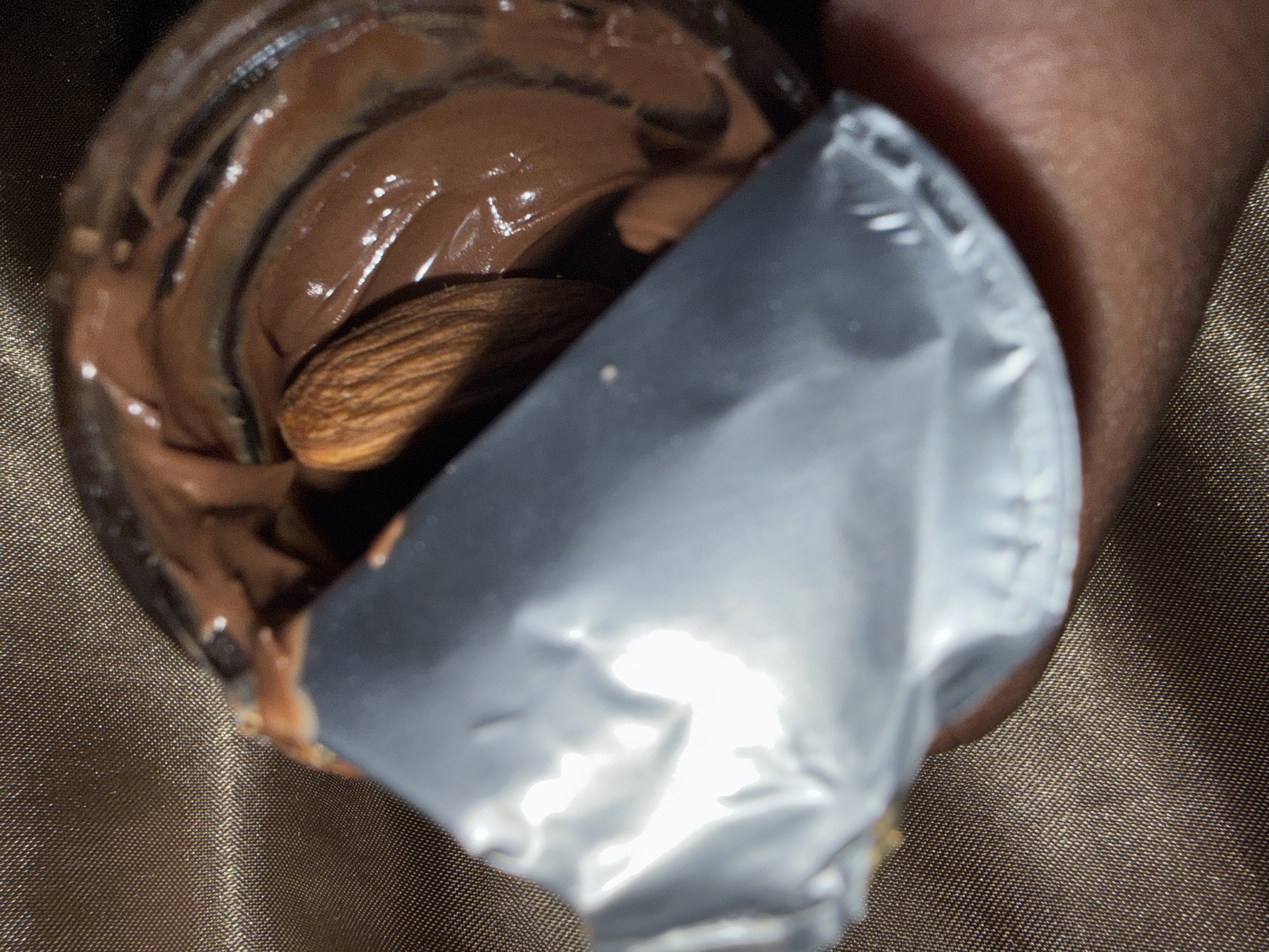 A close-up shot shows an open jar of chocolate spread with an almond visible within. The jar's foil lid is partially peeled back. The jar is held in someone's hand. The background appears to be a textured fabric. There is no indication in the image of Sarcelles, France.