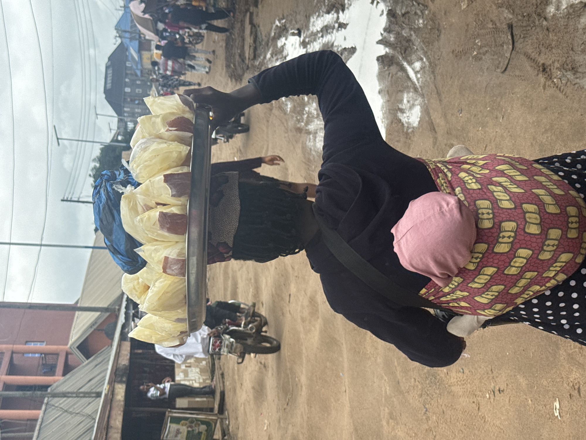 In the city of Ihiagwa, Nigeria, a woman carries a tray of food items wrapped in plastic bags on her head. She is also carrying a baby wrapped in a patterned cloth on her back. The woman is wearing a dark hooded top and a polka dot skirt. The ground is dusty and uneven, with puddles of muddy water visible. In the background, there are buildings, some with corrugated metal roofs, and other people walking. A motorcycle is also visible. The sky is overcast.