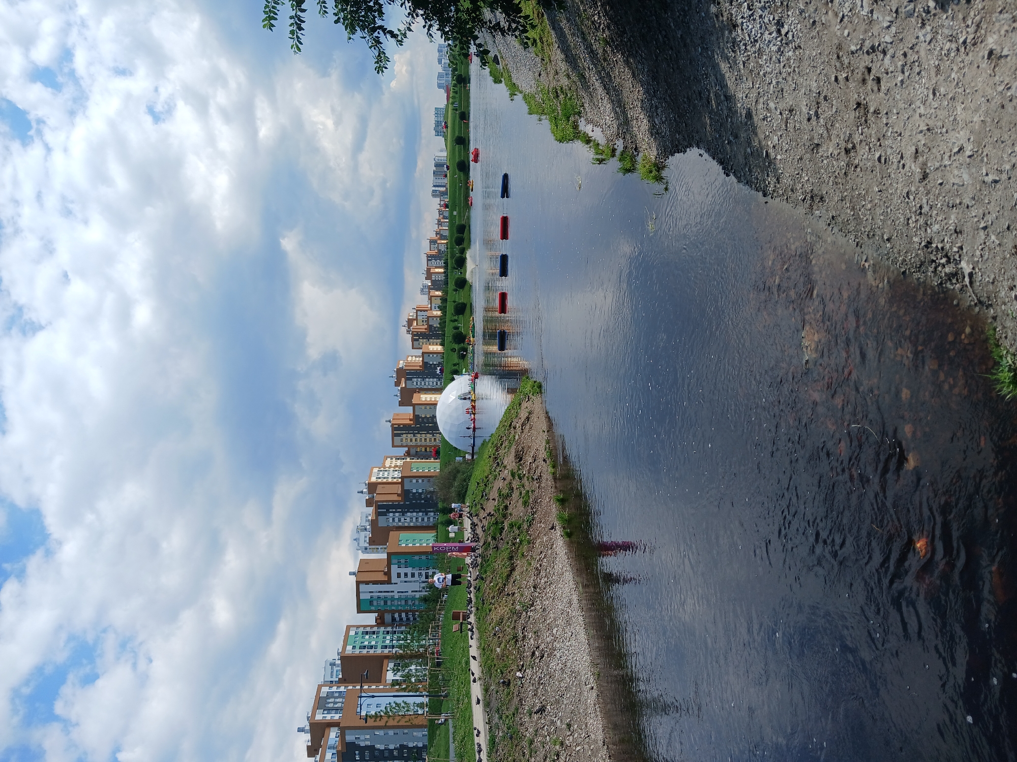 The image captures a scenic view of a body of water with a cityscape in the background, situated in Yekaterinburg, Russia. The foreground features a calm body of water with visible pebbles and rocks beneath the surface near the edge. The left bank of the water is a sloped, grassy area with a walking path. A man is standing on the path, facing the water, with a flock of pigeons gathered around him. Several other individuals, including children, are also seen walking on the path in the mid-ground. To the right, the bank is rocky and sparsely vegetated.
In the middle ground, across the water, a large, white dome-shaped inflatable structure is present. Surrounding this dome are several small boats, some of which are red and blue, floating on the water. In the background, a modern residential area with multi-story buildings of varying colors, including beige, green, and grey, stretches across the horizon. The sky is partly cloudy, with a mix of blue sky and white clouds. A tree branch with green leaves is visible in the upper right corner of the frame. The overall impression is that of a recreational area within a developed urban environment.