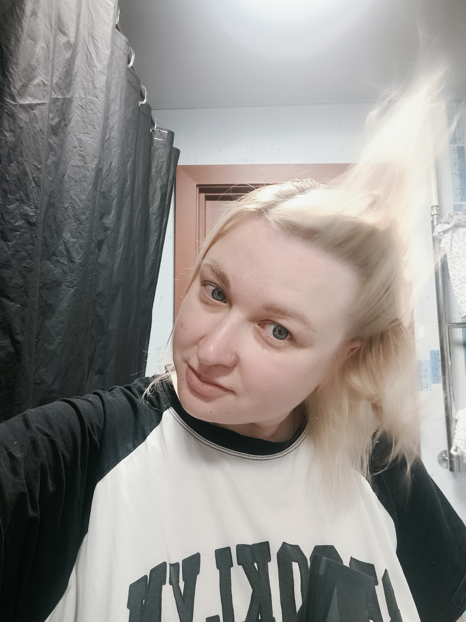 A selfie captures a young adult woman with light blonde hair and blue eyes, appearing to be in a bathroom in Chaikovsky, Russia. She looks directly at the camera with a subtle, friendly smirk. Her fair skin shows a few minor blemishes.
Her light blonde hair is prominently featured, styled or caught in motion, extending upwards and to the right, appearing brightened, possibly by a flash or a light source from a device she might be holding. She is wearing a casual black and white raglan-style t-shirt with black sleeves and a white body. Part of a dark text graphic, possibly "BROOKLYN," is visible on the white section of her shirt, largely obscured by a black object that resembles part of a hairdryer.
The background elements clearly identify the setting as a bathroom. To her left, a dark grey or black shower curtain hangs from a rail with white rings. To her right, a light brown door frame is visible next to a wall covered in light blue tiles. Further right, a mirrored cabinet or shelf, adorned with a white patterned towel, can be seen. The ceiling is white, and the lighting is bright and even, typical of indoor artificial light. The image provides a close-up view of the woman's face and upper body, with the bathroom details framing her.