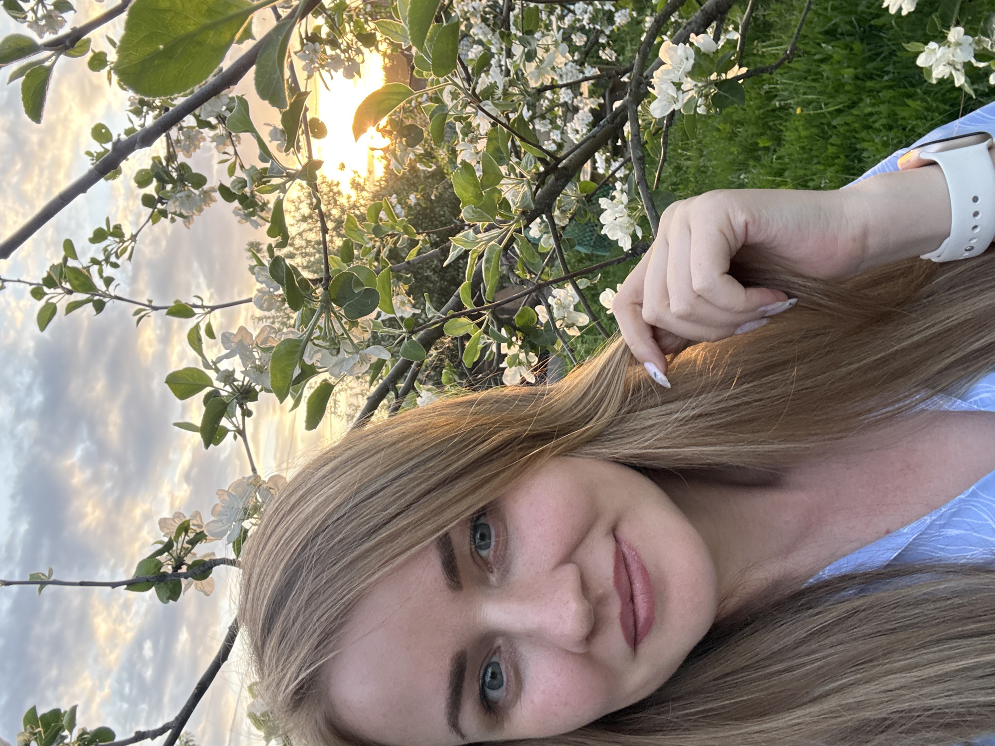 A medium shot of a blonde woman with blue eyes, looking into the camera, smiling. She is in an outdoor setting with blooming trees and a sunset in the background. She is wearing a light blue shirt and a white smartwatch. The location appears to be Chaikovsky, Russia, based on visual elements.