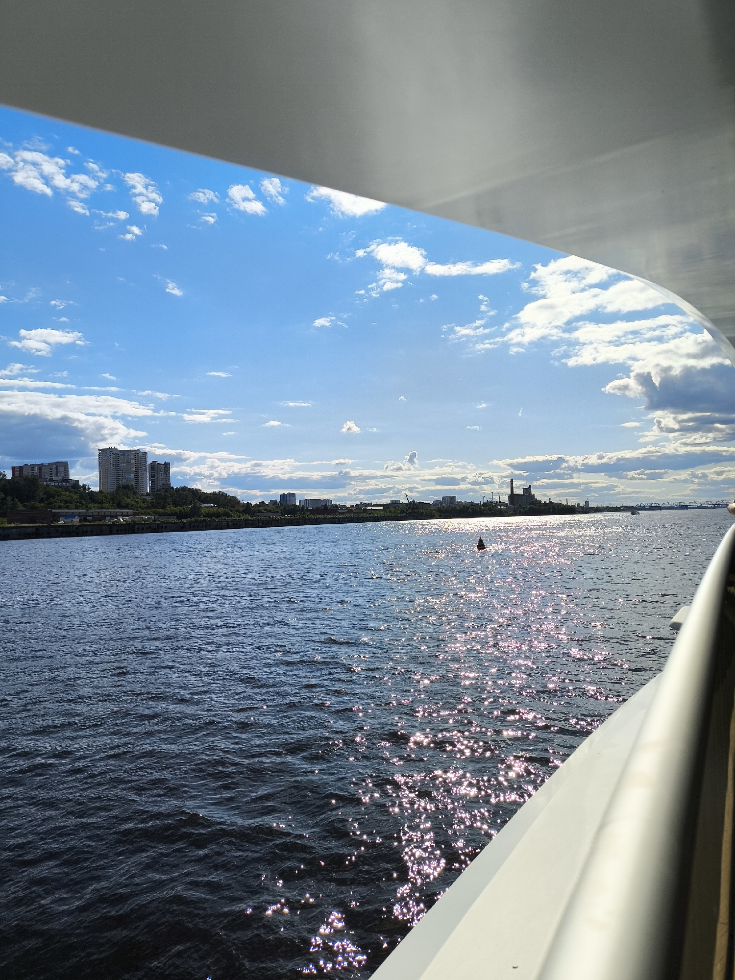 This is a photograph taken from a vessel on the Kama River in Perm, Russia, looking towards the left bank under a bright blue sky with scattered white clouds. The sun reflects off the water, creating a shimmering effect. In the distance, the Perm skyline is visible, featuring several multi-story apartment buildings, industrial structures including a power plant with a smokestack, cranes, and a bridge. A red buoy is present in the foreground of the water. The edge of the vessel from which the photo is taken is visible in the lower right corner, with a white railing.