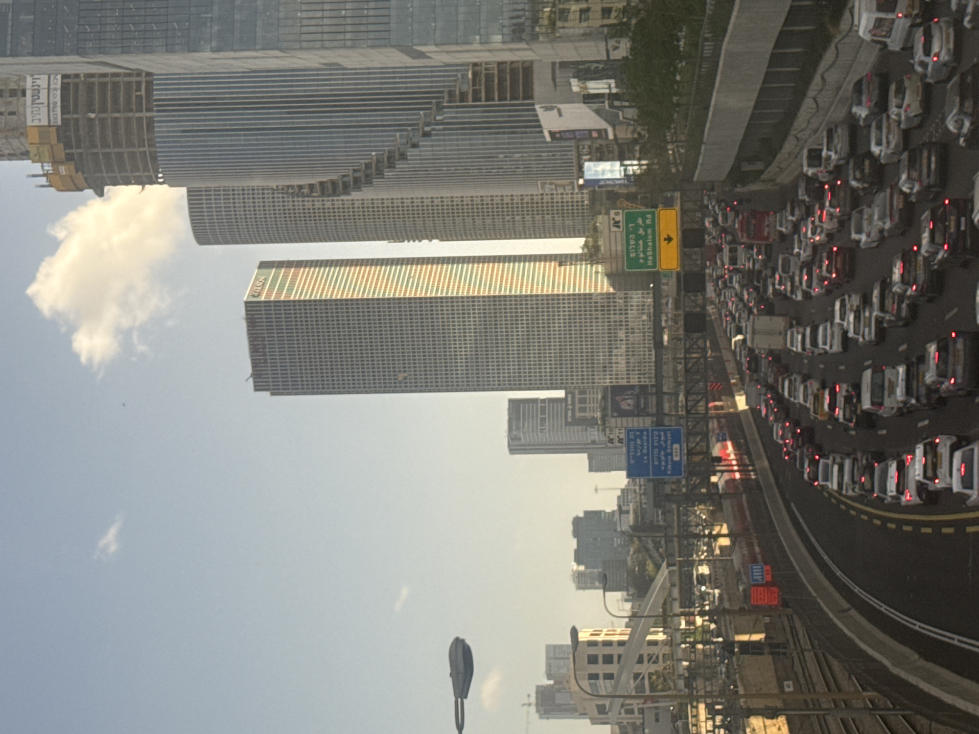 The image shows a view in Tel Aviv, Israel, featuring high-rise buildings, including the Azrieli Center, against a sky with a cloud. A busy highway is filled with cars, with their brake lights visible. Road signs indicating directions to HaShalom Road and other locations are present. Streetlights and various urban structures are part of the cityscape.