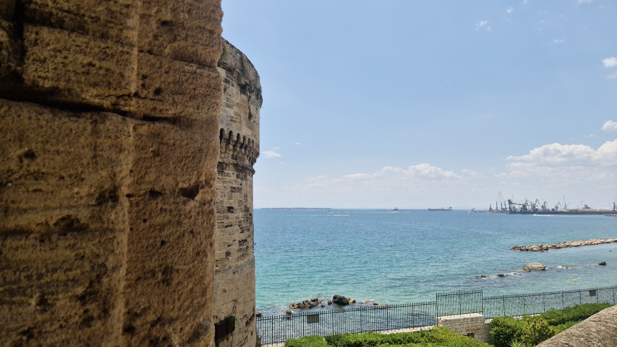 The image shows a view from a stone structure overlooking the sea. In the foreground, a large, rough-hewn stone pillar occupies the left side of the frame, textured with visible pores and embedded with smaller stones. To the right of the pillar, a portion of a weathered stone wall or fortification extends into the frame, featuring arched openings and rough stonework.
Beyond the fortification, a body of turquoise-colored water stretches to the horizon. The water appears calm with gentle ripples. Along the coastline in the distance, a port or industrial area is visible, characterized by numerous cranes, ships, and structures. The sky is a clear blue with scattered white clouds. A thin line of land or a breakwater extends into the sea on the right side of the frame, with rocks and vegetation along its edge. A black metal fence runs along the top of a low stone wall in the mid-ground, separating the viewer's immediate location from the sea. The overall scene suggests a historical site with a view of a working port. This view is likely from the Aragonese Castle (Castello Aragonese) in Taranto, Italy, which offers panoramic views of the Mar Piccolo and the port area. There are no people or animals visible in the image.