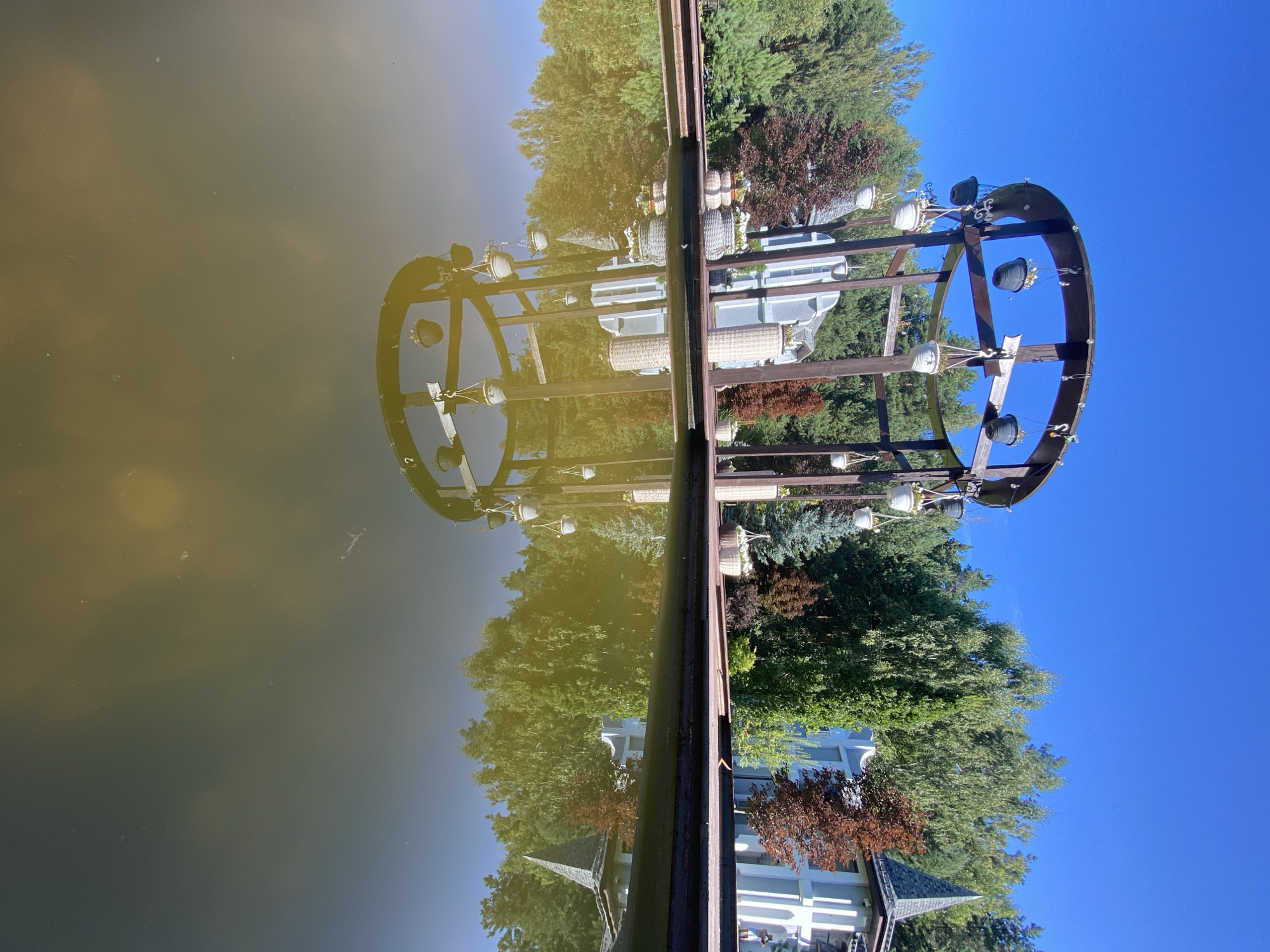 A low-angle, full outdoor shot captures a serene scene at a pond in Chulis, Russia, on a clear, sunny day. The water's surface reflects the clear blue sky and a wooden gazebo adorned with hanging flower baskets, as well as trees and a building with a pointed roof. The gazebo, made of dark wood, has a circular top and vertical supports. White and dark pots containing flowers hang from the gazebo's structure. Beyond the gazebo, lush green trees form a backdrop, with a building of white and blue architecture featuring a prominent pointed roof. The foreground is dominated by the pond and its reflections, with the wooden deck surrounding the pond visible along the water's edge. No people are clearly visible in the frame.