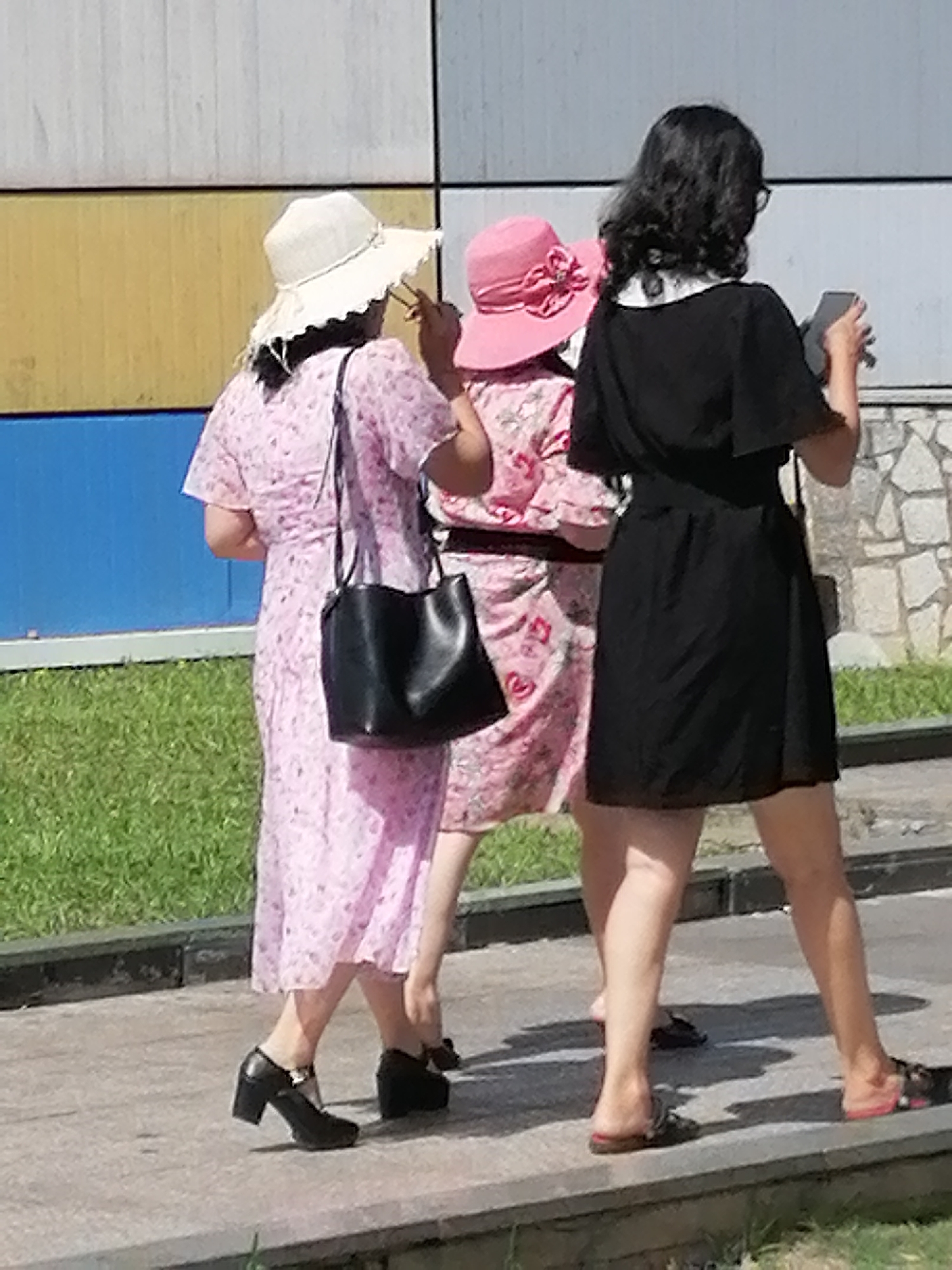 Three individuals, appearing to be women, are walking outdoors, possibly in Da Lat, Vietnam. The first individual from the left is wearing a light-colored wide-brimmed hat and a pink floral dress. She carries a black handbag on her left shoulder. The second individual, in the middle, is wearing a pink hat with a decorative bow and a floral dress. The third individual on the right is wearing glasses and a black dress. She is holding a smartphone in her right hand and appears to be taking a photo or using the device. They are walking on a paved walkway with a grassy area to their left and a building with a textured wall to their right. The lighting suggests it is daytime, with shadows cast on the ground.