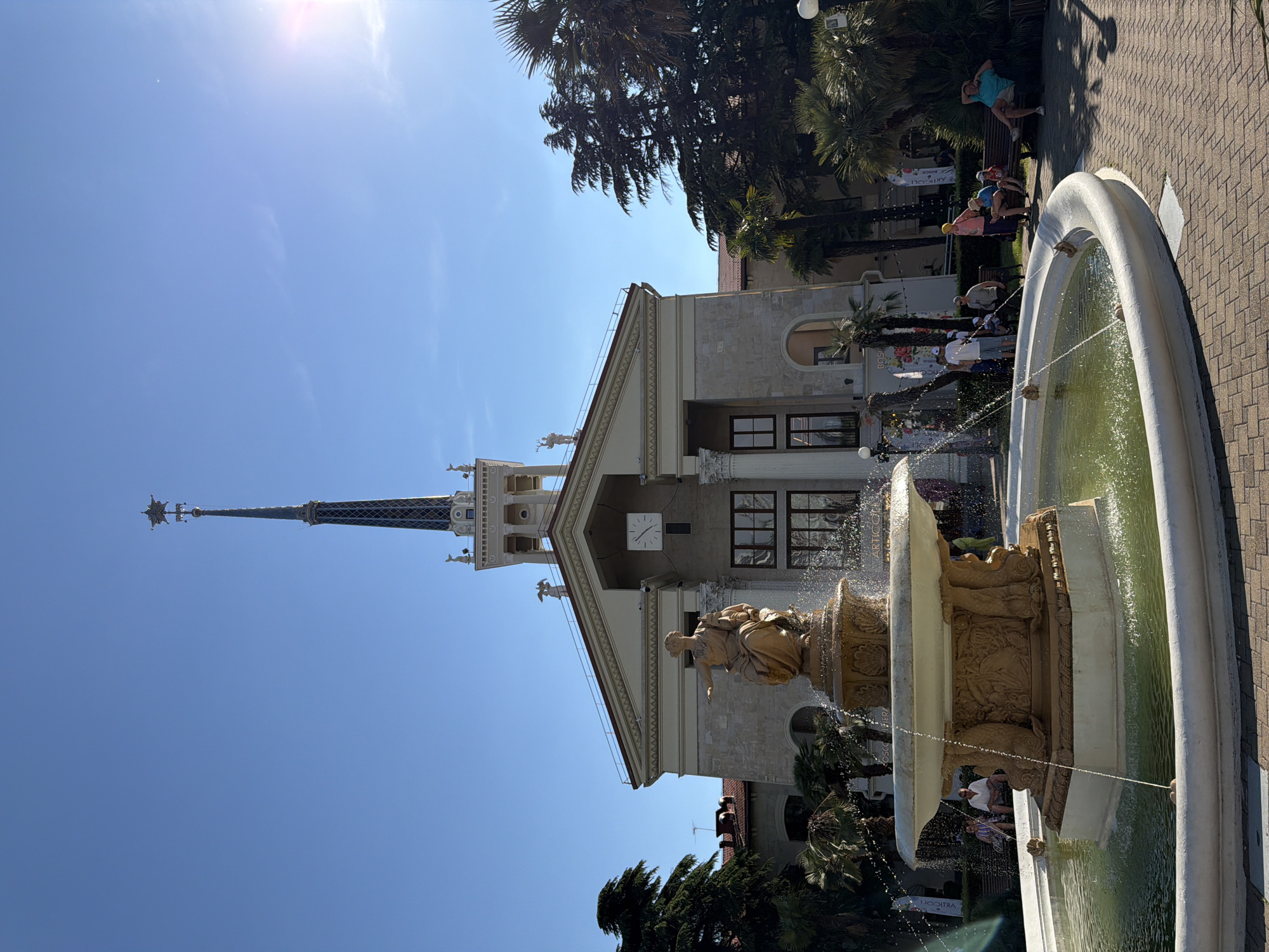 The image captures a sunny day in Sochi, Russia, with a clear blue sky dominating the frame. The scene is a lively outdoor public space featuring a grand fountain in the foreground, its water spraying upwards. The fountain is adorned with ornate sculptures. Behind the fountain, a building with classical architectural elements stands tall, characterized by a prominent clock on its facade and a pointed spire reaching towards the sky. Palm trees and other lush greenery are scattered throughout the scene, adding to the tropical feel.
Several people are visible in the image, some seated on benches and others standing near the fountain. Their attire suggests warm weather and a relaxed atmosphere, with individuals casually dressed. A few people are captured in mid-action, possibly posing or enjoying the ambiance. The overall mood appears to be cheerful and leisurely, indicative of a popular tourist or recreational spot.
The building's design, with its decorative elements and spire, suggests it might be a historical landmark or a significant public structure. The presence of palm trees strongly points towards a coastal or subtropical region, which aligns with the given location context of Sochi, Russia. The bright sunlight and clear skies indicate it is daytime, likely mid-morning or afternoon.
While most of the text in the image is illegible due to distance and resolution, a partially visible sign with Cyrillic lettering can be seen on a banner in the background. The fountain itself is a focal point, with its intricate design and the dynamic spray of water creating a sense of movement and vibrancy.