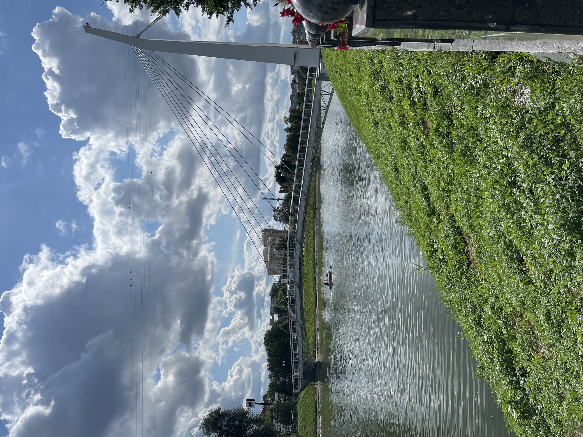 The image captures a view of the Staryi Saltiv River in Kharkiv, Ukraine, on a cloudy summer day. A modern pedestrian bridge, characterized by its white pylon and tension cables, spans the river. On the water, a small boat with at least two individuals can be seen paddling. The riverbank on the right side of the image is covered in lush green grass, with a dark stone railing and decorative spherical elements in the foreground. The opposite bank features trees, buildings, and utility poles. The sky is filled with dramatic cumulus clouds, interspersed with patches of blue. In the upper portion of the image, several birds are perched on power lines.