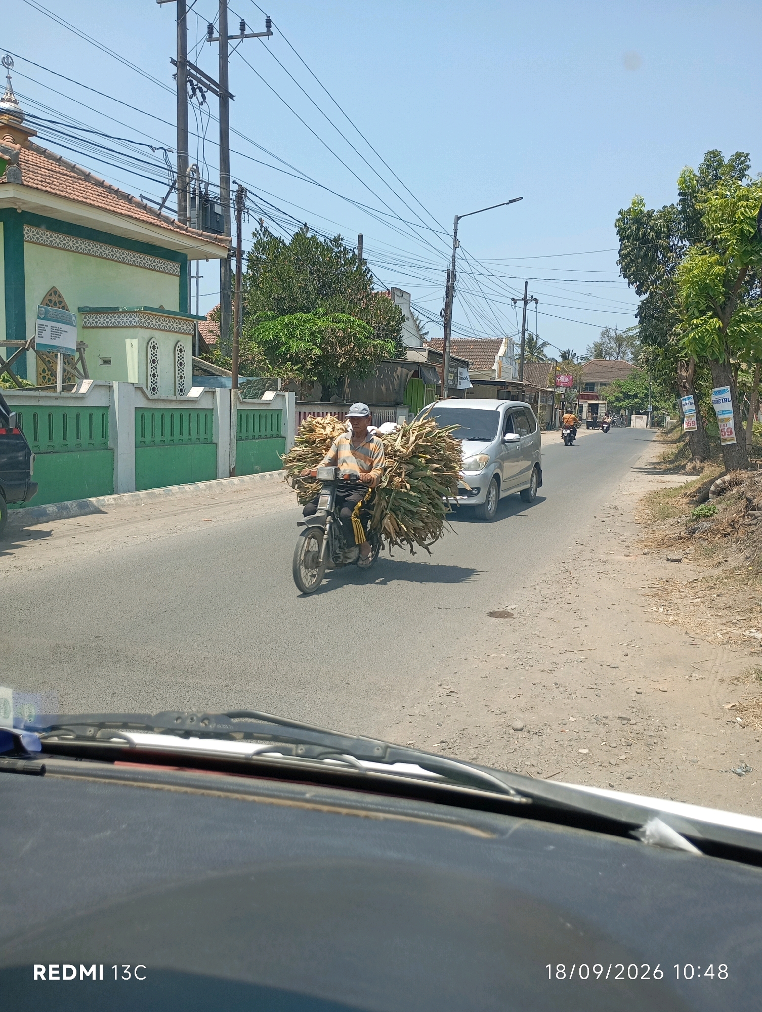 The image depicts a bright, sunny day in Sumberrejo, Indonesia, on a paved road. In the foreground, the dashboard of a vehicle with windshield wipers is visible, with a timestamp "18/09/2026 10:48" and "REDMI 13C" in the lower right and left corners respectively. A man, wearing a light-colored, horizontally striped shirt and a blue cap, is riding a motorcycle loaded with a large bundle of corn stalks. He is traveling on the road, with a silver minivan to his left and other motorcycles further down the road. To the left of the road, a light green building with a tiled roof, possibly a mosque due to its architectural features and crescent moon symbol, is visible, bordered by a green fence. The environment features utility poles with numerous wires, trees with green foliage, and a dusty roadside.