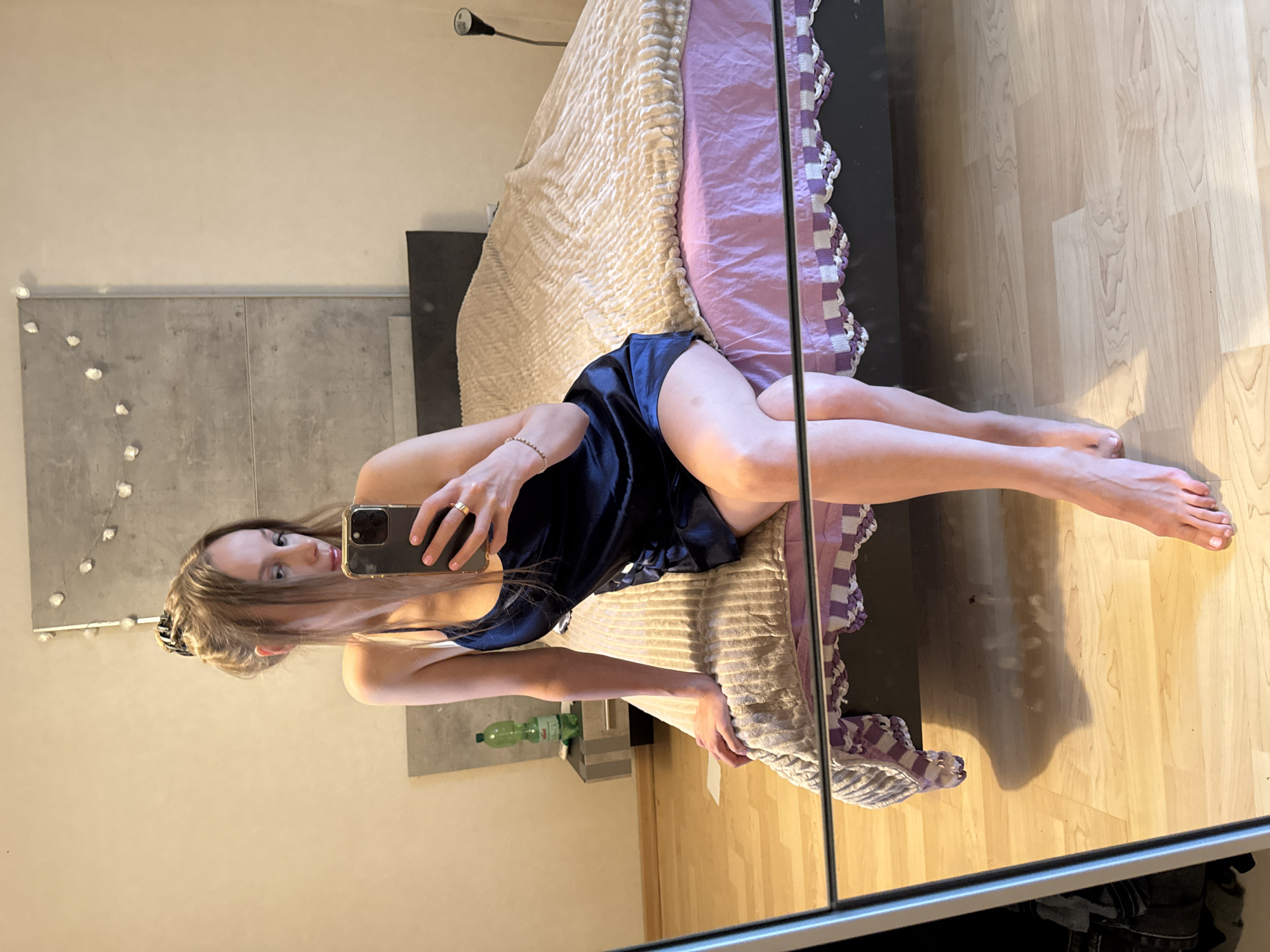 A young woman is taking a mirror selfie in a bedroom. She is sitting on the edge of a bed, wearing a dark blue slip dress. Her hair is pulled up in a messy bun. She is holding a phone in her right hand and a ring is visible on her left ring finger. The bed has a light brown textured comforter and a purple sheet with a white and purple trim. A bedside table with a water bottle is visible to the left of the bed. The room has light-colored walls and a wooden floor. A large mirror reflects the scene, showing the woman and a portion of the room. The overall impression is that of a casual, private moment. The location of Überherrn, Germany, is not discernable from the image itself.