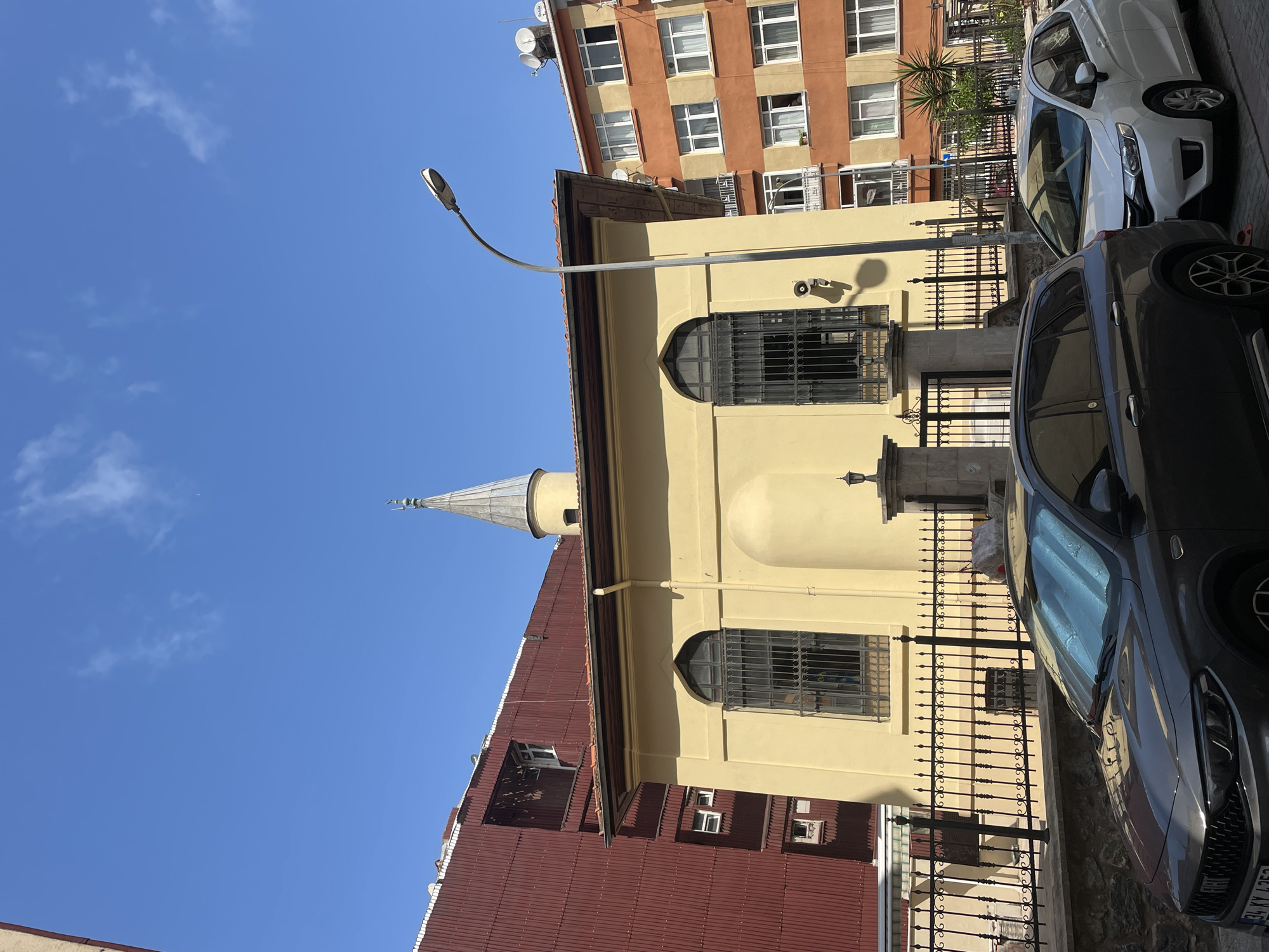 A low-angle, full shot captures a yellow mosque with a conical minaret against a clear blue sky with sparse clouds. The mosque features arched windows with dark metal grills. To the left of the mosque, a tall, red corrugated metal building stands, with several windows visible. To the right of the mosque, a multi-story orange and beige apartment building is present, also with numerous windows. In the foreground, two cars are parked on a street: a dark gray SUV on the left and a white sedan on the right. A black metal fence with decorative elements runs along the front of the mosque's property. The street is paved, and a streetlamp is visible on the right side of the image. The image appears to be taken during daylight hours. The scene is located in Kadıköy, Turkey.