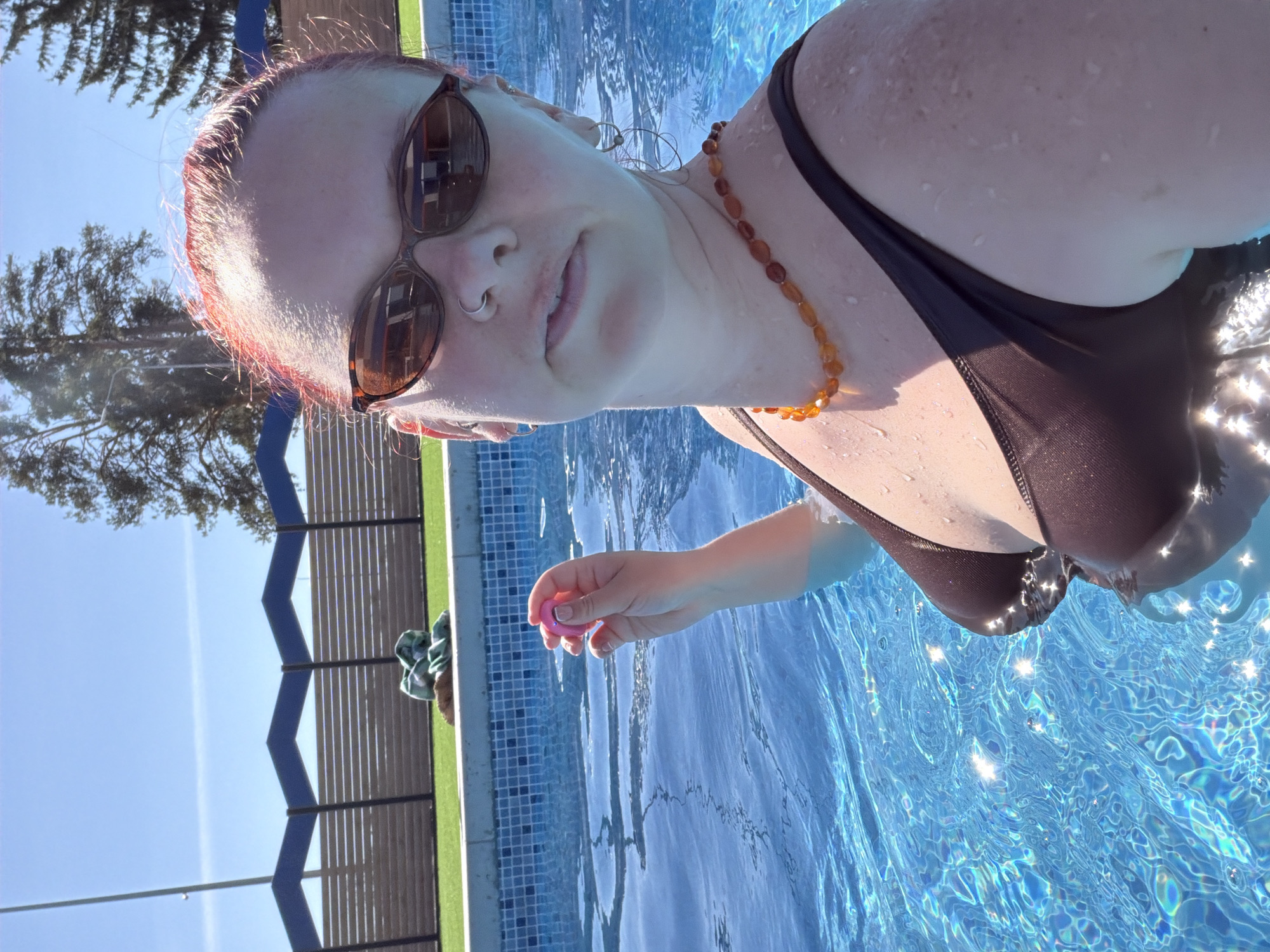 A woman is swimming in a pool. She is wearing sunglasses, a dark-colored swimsuit, and a beaded necklace. She has pink-tinted hair. She is holding a pink object in her right hand. The pool is surrounded by a fence and green turf. The location is likely a private pool and there is no way to determine if it is located in Votkinsk, Russia from this image.