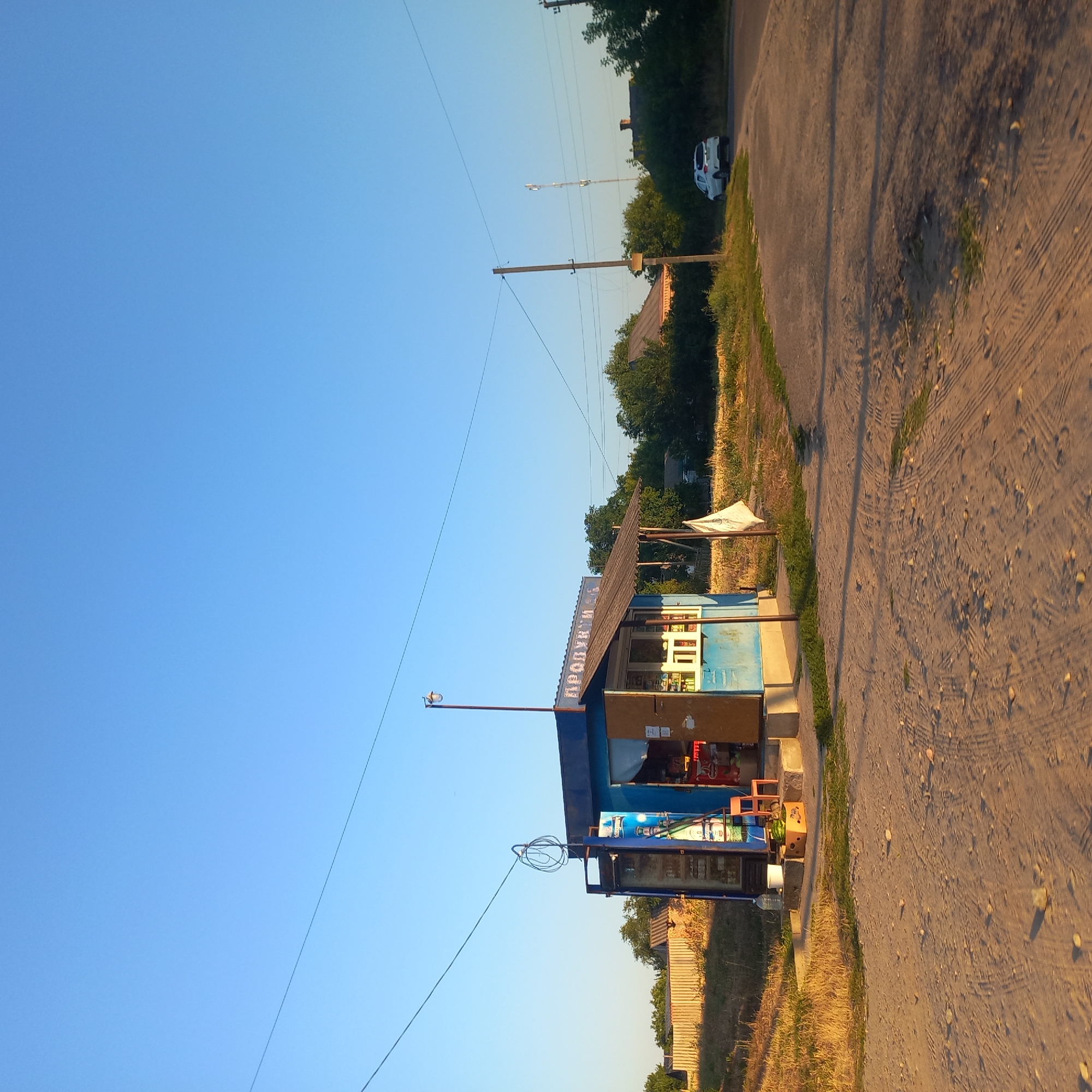 A small, blue, open-air kiosk, labeled "ПРОДУКТИ" (Products), is situated on a dusty, unpaved road in a rural area, possibly on the outskirts of Klyntsi, Ukraine. The kiosk has a wooden counter and display shelves visible, with various products stocked inside. A small orange chair is placed near the entrance. In front of the kiosk, there's a cardboard box containing watermelons and a large plastic bottle of water. To the left, a wooden fence borders a grassy area. A white flag on a pole stands to the right of the kiosk. In the background, trees and some houses are visible. A white car is parked on the side of a paved road further in the distance, and a cellular tower can be seen on the horizon. The sky is clear and blue, indicating daytime. The ground is a mix of dirt, sand, and sparse vegetation. Power lines are strung across the scene.