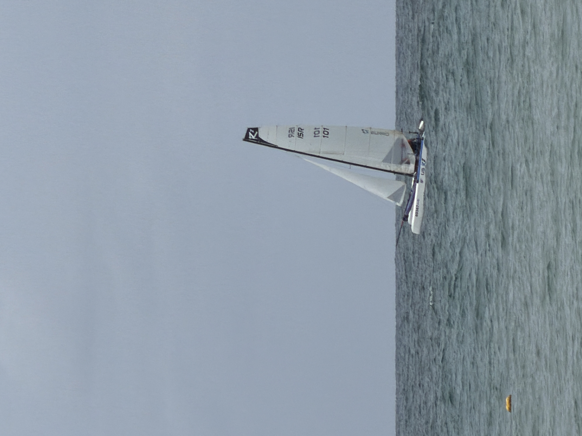The image depicts a sailboat on the water, with the sky filling the left side of the frame. The sailboat has white sails with the numbers "101", "ISR", and "121" visible. The ocean water is gray and choppy. A small, yellow object appears in the water near the bottom right corner. The scene appears to be in Tel-Aviv, Israel due to the sail registration number, ISR.