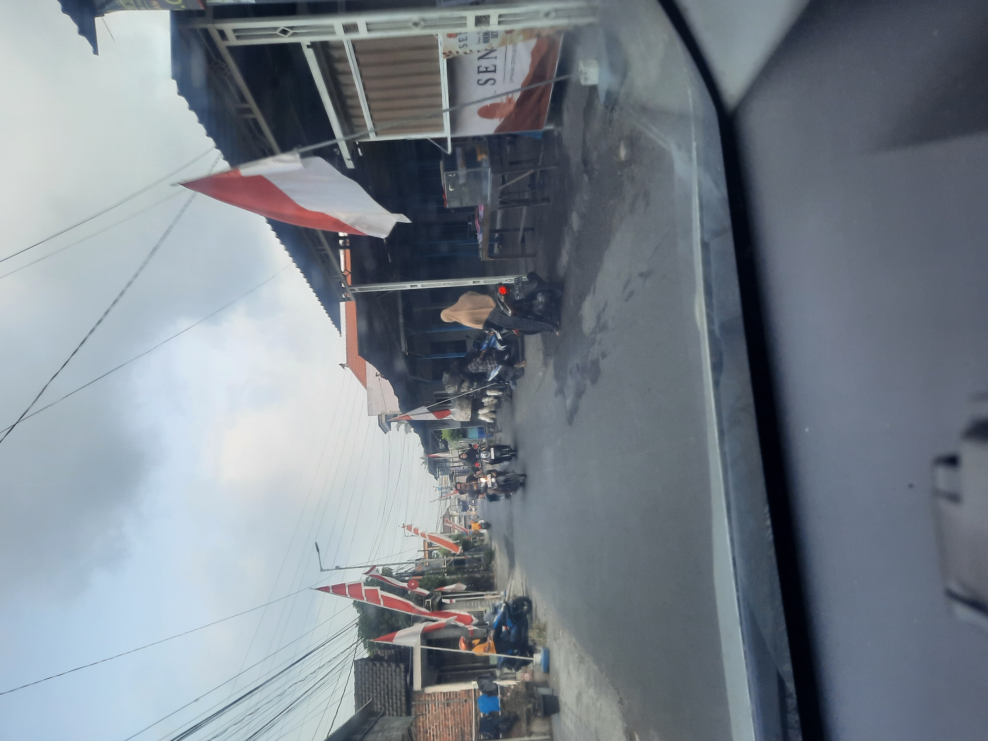 The image shows a street scene in Sidokepung, Indonesia. The road is lined with buildings, some of which appear to be shops or market stalls. Indonesian flags, red and white, are prominently displayed along the street, suggesting a national celebration or commemoration. Several people are present, mostly on motorcycles, navigating the road. One person, wearing a beige headscarf, is seated on a motorcycle. Another individual in a yellow shirt is also on a motorcycle. The sky is overcast with clouds. The foreground shows the interior of a vehicle, likely from which the photo was taken, with a dashboard and windshield visible.