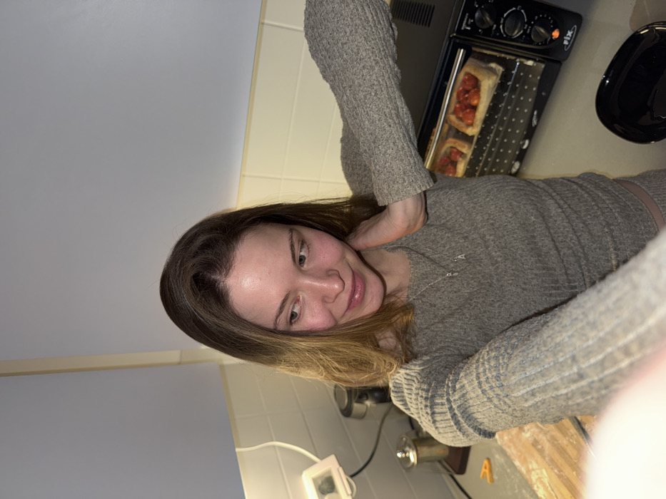 The image depicts a domestic kitchen scene in Tel-Aviv, Israel. A light-skinned female subject with medium-length brown hair occupies the foreground, extending her left arm towards the camera, indicating a self-portrait. She wears a light brown, ribbed long-sleeved top and a silver necklace with an 'A' pendant. Her right hand rests on the back of her neck/shoulder, and her gaze is directed slightly to her left (viewer's right) with a subtle smile.
The background reveals a kitchen countertop with a light yellow-beige tiled backsplash and light-colored overhead cabinets. On the countertop to the subject's right, a black "Rix" brand mini-oven is visible, with an orange indicator light illuminated, indicating operation. Inside the oven, two rectangular pastry items topped with small red elements, consistent with cherry tomatoes, are baking. To the subject's left, a white power outlet, a small white digital device, a partial silver kettle, and a metallic cylindrical container are visible. A flour-dusted wooden cutting board with a small orange plastic clip is present in the lower-left foreground, and a black bowl is on the countertop in the lower-right. The scene is illuminated by artificial lighting.