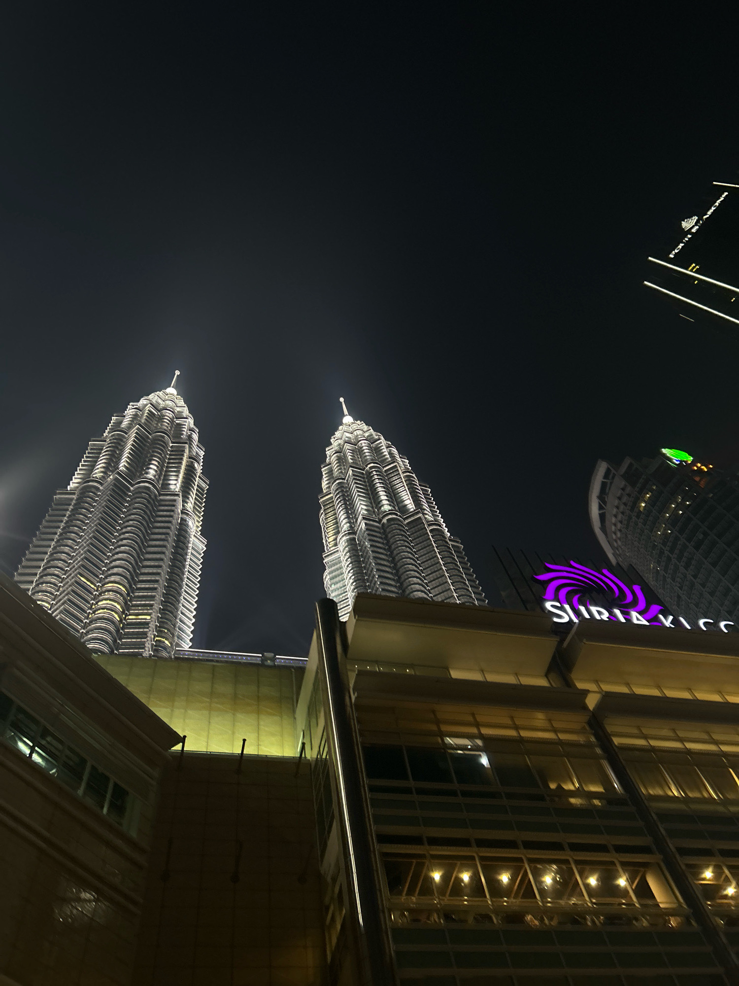 This image captures the Petronas Twin Towers in Kuala Lumpur, Malaysia at night. The iconic skyscrapers, illuminated by white lights, dominate the upper portion of the frame. In the foreground, a modern building with large glass windows and illuminated interiors is visible. The building also features a prominent sign with purple lettering that reads "SURIA KICC." The sky is dark, indicating it is nighttime. No people or vehicles are clearly discernible in the image. The overall scene depicts an urban nightscape with prominent architectural landmarks.