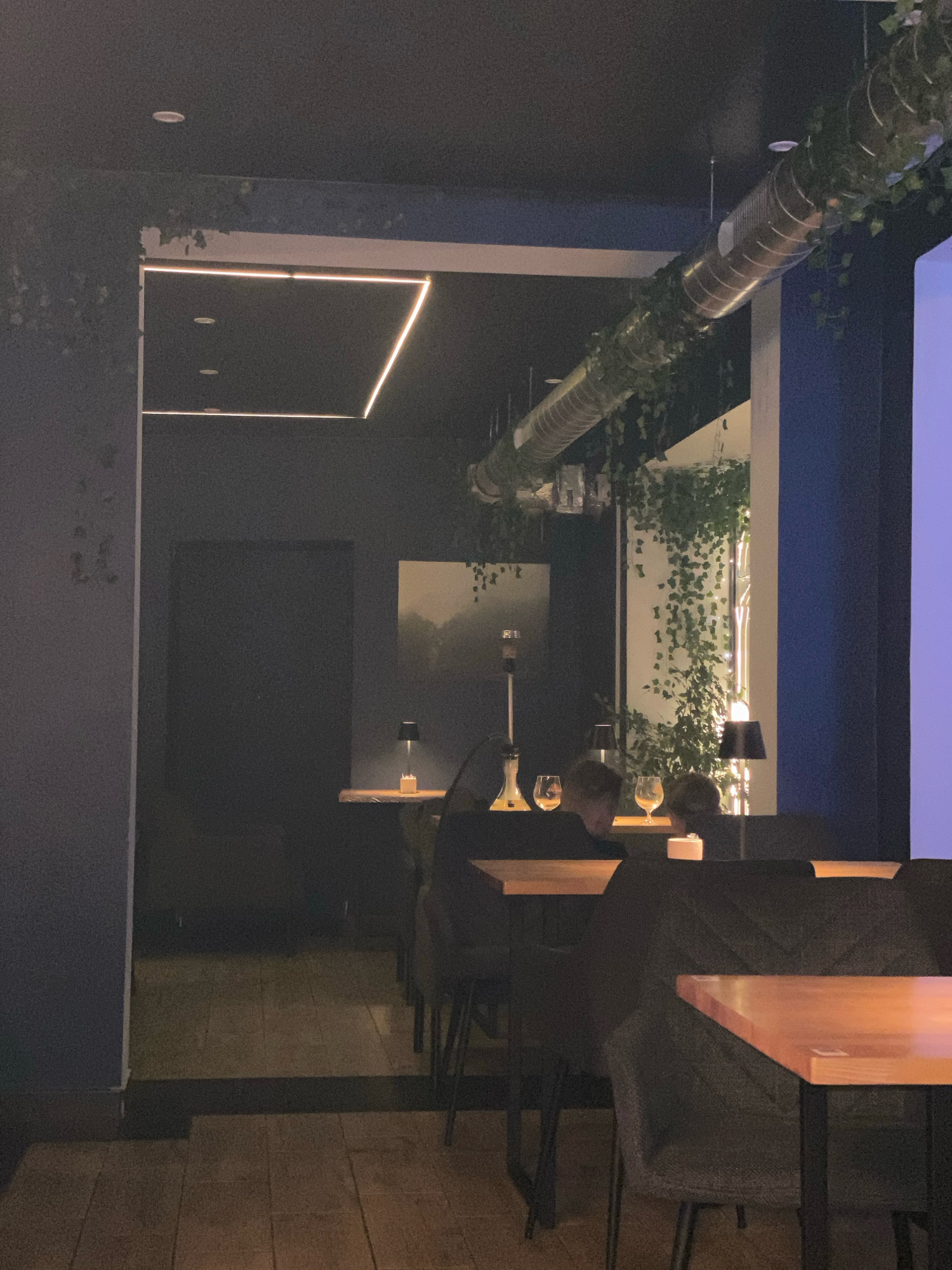 The image depicts the interior of a dimly lit establishment, likely a bar or lounge, with a modern industrial aesthetic. Dark walls and a dark ceiling are punctuated by geometric LED lighting. A large, corrugated metal ventilation duct, adorned with artificial greenery, runs across the upper portion of the scene.
In the foreground, several tables and chairs are visible. One table in the lower right has a light-colored wooden top and dark legs, with a dark upholstered chair partially visible. To the left, another table has a similar wooden top and dark legs, with two chairs in front of it. A hookah pipe stands on a table in the middle ground, with two wine glasses placed nearby. The presence of a hookah suggests the venue may be a shisha bar.
In the background, a doorway leads to a darker space with a wooden floor. A framed artwork, depicting a misty landscape, hangs on a dark wall. Small table lamps with black shades are placed on shelves, casting a warm glow. The overall ambiance is moody and intimate, with artificial plants contributing to a biophilic design element.
There is no visible information within the image to ascertain its specific location in Gliwice, Poland.