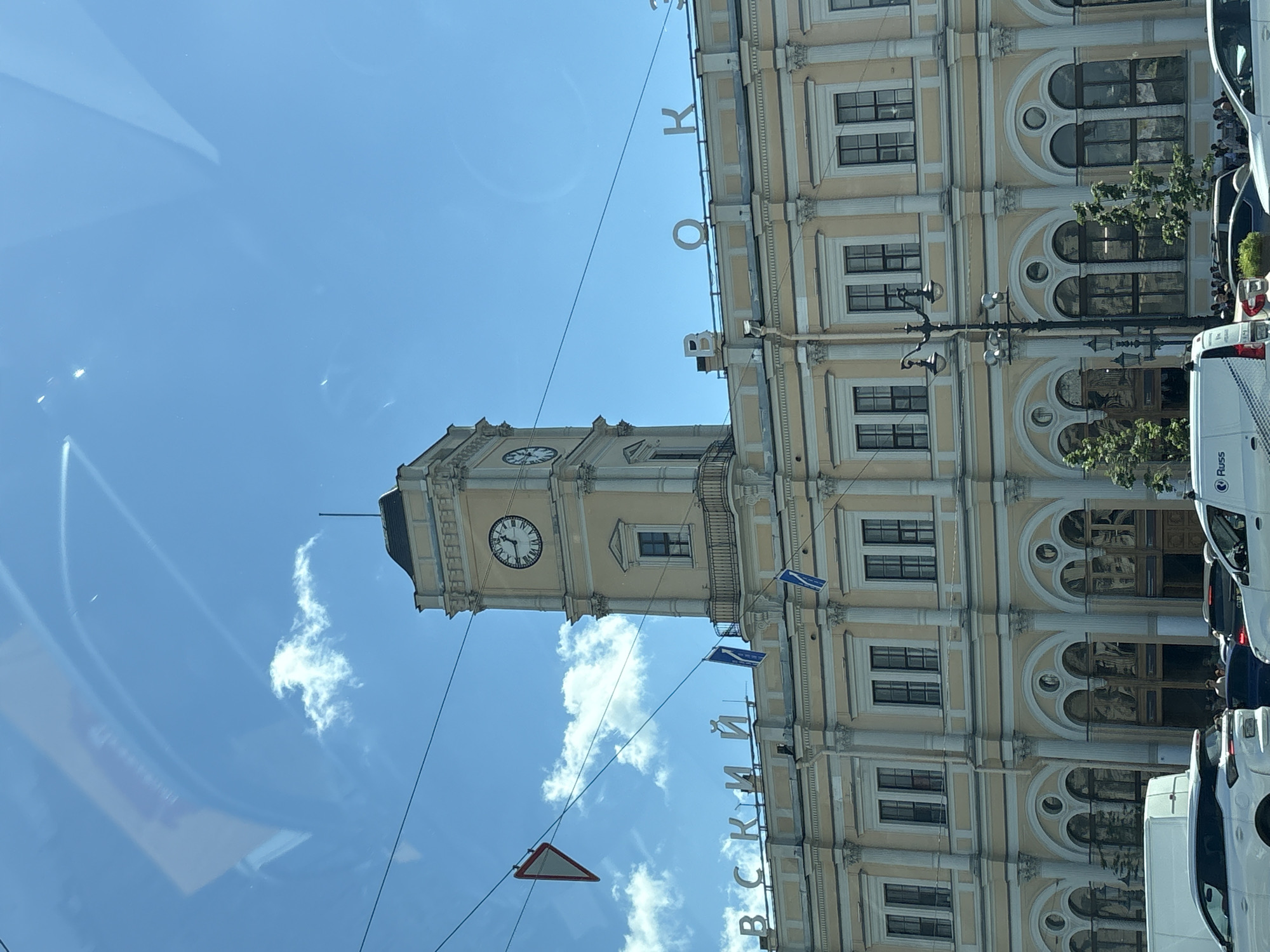The image shows the facade of a train station in Saint Petersburg, Russia, likely the Vitebsky Railway Station due to the Cyrillic lettering visible on the building. The station is a grand, light yellow building with classical architectural elements, including columns and arched windows. A prominent clock tower rises above the main structure, featuring Roman numerals on its clock face. In front of the station, there is a busy street with several cars and a white van. The sky is blue with some scattered clouds, and power lines are visible crisscrossing the frame. The scene appears to be captured from a moving vehicle, as evidenced by reflections on the glass in the foreground.