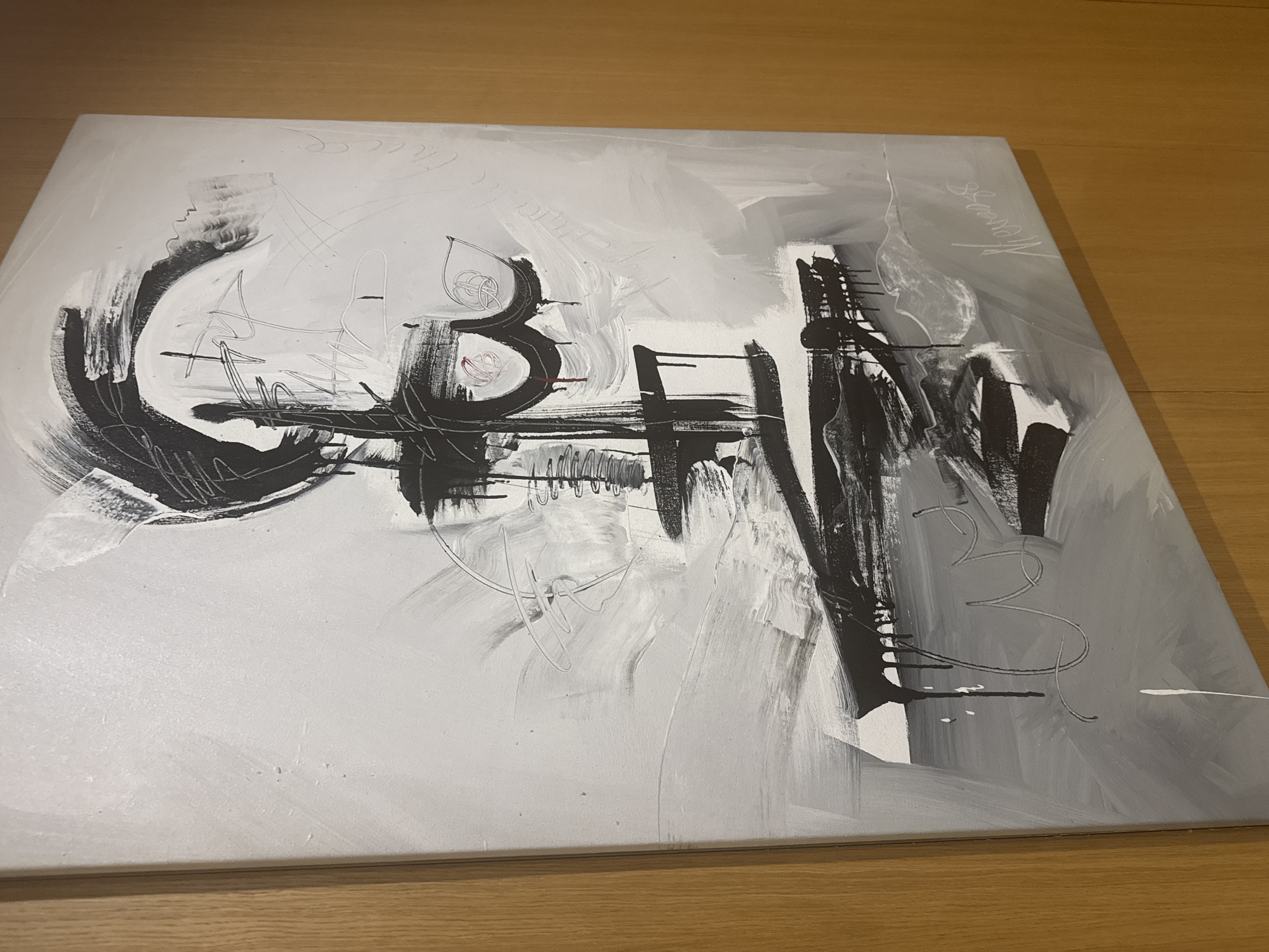 The image shows a close-up, angled view of an abstract painting with black and white brushstrokes, accented by drips and some light grey and white textures. The painting is framed by a light-colored wooden border on the left and right sides, suggesting it is hanging on a wall in a room. The background wall appears to be a warm, light brown, possibly wood paneling. No specific location in Barcelona, Spain, can be identified from this image.