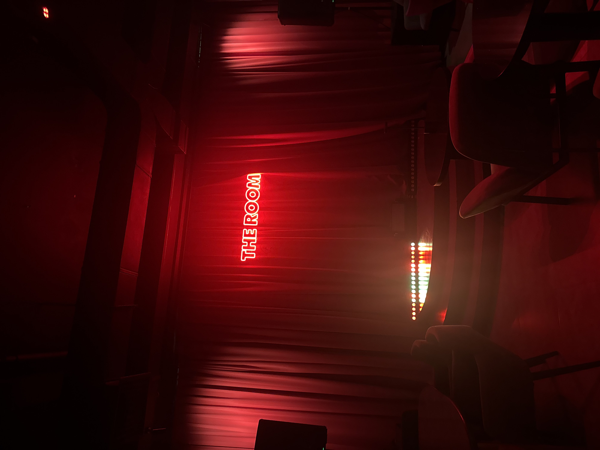 The image depicts an indoor scene with a red neon sign that reads "THE ROOM" prominently displayed against a backdrop of red velvet curtains. The space appears to be a stage or performance area, indicated by a raised platform in front of the curtains, illuminated by a line of colorful stage lights. The overall ambiance is dimly lit with a strong red hue, suggesting a bar, club, or intimate performance venue. In the foreground, several chairs and tables are visible, some casting long shadows, further emphasizing the low lighting conditions. The location is identified as North Kuta, Indonesia. There are no people clearly visible in the frame, and the scene appears to be staged for an event or performance. The architecture includes exposed ceiling beams and ventilation systems, hinting at a possibly industrial or modern design.