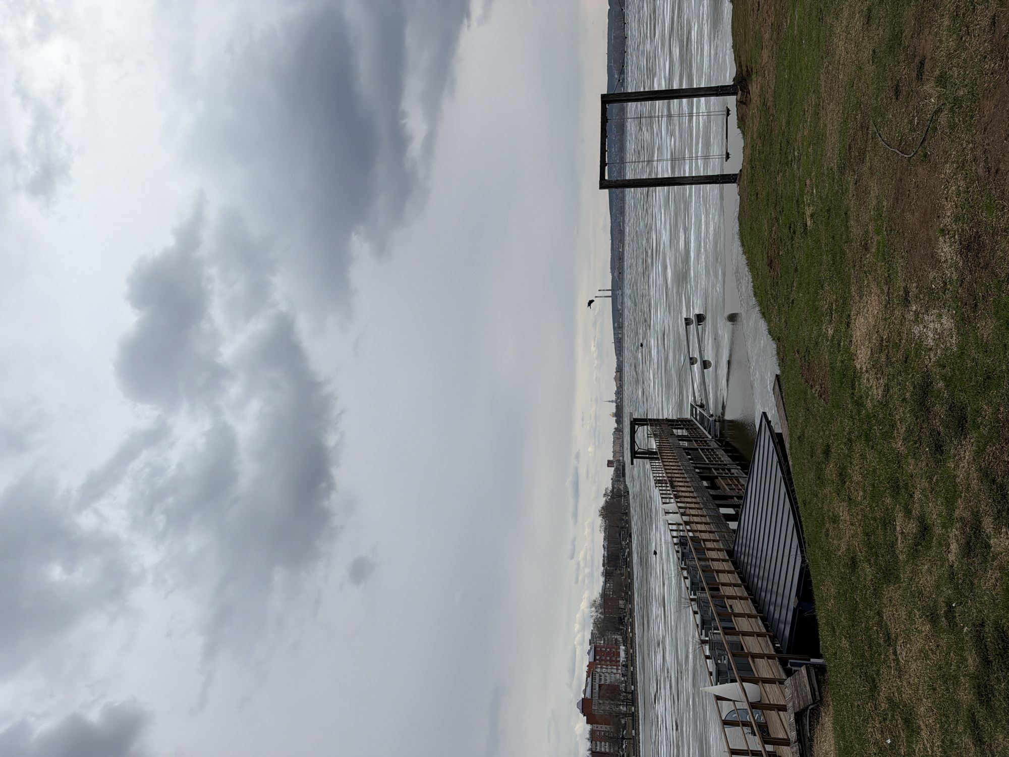 The image shows a riverfront scene in Votkinsk, Russia, on an overcast day. The view is oriented with the river running horizontally across the frame. On the right, there is a grassy bank. A wooden pier extends out into the river. The river appears choppy or potentially frozen. In the distance, the city skyline is visible along the opposite bank of the river. The sky is heavily clouded. A rectangular metal structure stands at the edge of the water near the grassy bank. A power line runs along the grass on the right. The image is shot from a slightly elevated position.
