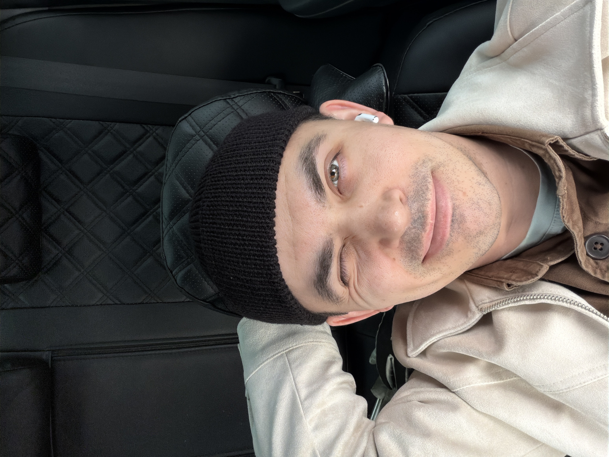 A man wearing a black beanie, a tan suede jacket over a brown collared shirt and a white earbud in his right ear is lying down on a black leather car seat with a quilted diamond pattern. The location cannot be determined from the image.