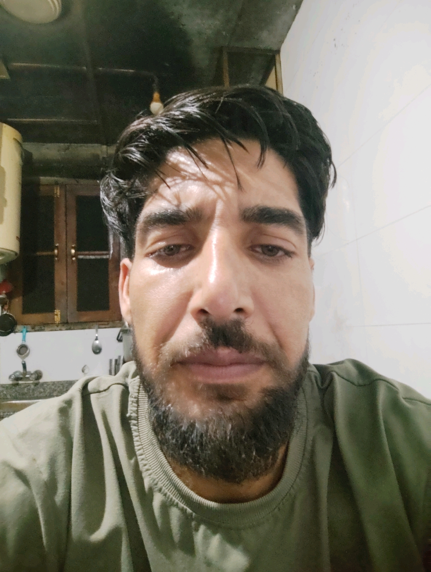 The image is a close-up, selfie-style photograph of a man's face and upper torso. The man has dark, wet-looking hair, dark eyebrows, and a beard. His skin appears slightly oily and is illuminated by a shadow pattern that suggests artificial lighting from above and to the left. He is wearing a muted green sweatshirt. The background shows a wall with some textured tiling or finish, and to the left, there is a wooden cabinet with two doors, a faucet with a spray attachment, and what appears to be kitchen or utility area elements. The overall environment suggests an indoor setting, possibly a home or a functional space. The image is not geographically specific to Srinagar, India, and no elements within the frame indicate such a location.