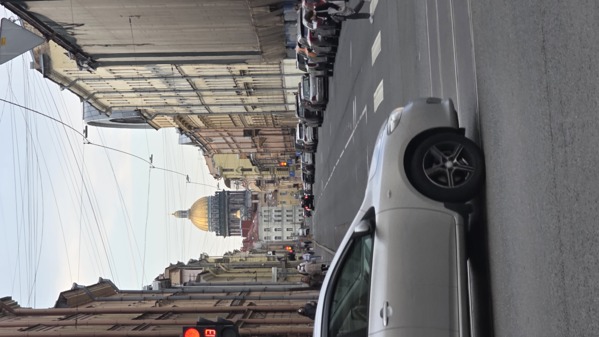 The image shows a street scene in Saint Petersburg, Russia, featuring Saint Isaac's Cathedral in the background with its large golden dome. The perspective is from street level, looking down a relatively narrow street lined with buildings of varying heights and architectural styles. The street is filled with parked and moving vehicles, including cars and what appears to be a motorcycle. Pedestrians are visible on the sidewalks, suggesting typical urban activity. The sky is overcast. The presence of overhead wires is noticeable, strung between the buildings. A car is parked on the right side of the frame, partially obscuring the street. Traffic lights are visible on the street.