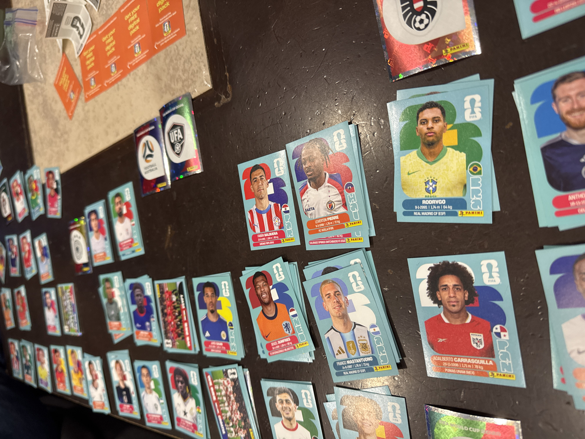 The image shows a collection of Panini FIFA World Cup 2026 sticker cards spread out on a dark, textured surface. The cards feature various soccer players and national teams. Some cards are stacked, obscuring the images beneath. Several are identifiable including those of players Rodrygo, Denzel Dumfries, Franco Mastantuono, and Adalberto Carrasquilla, as well as the logos of Football Australia and UFA. Paper promotional materials with text about "Get your FREE digital pack" are visible in the upper left corner, alongside a small plastic bag. There is nothing in the image to indicate a location in the city of San Bernardino Tlaxcalancingo, Mexico.