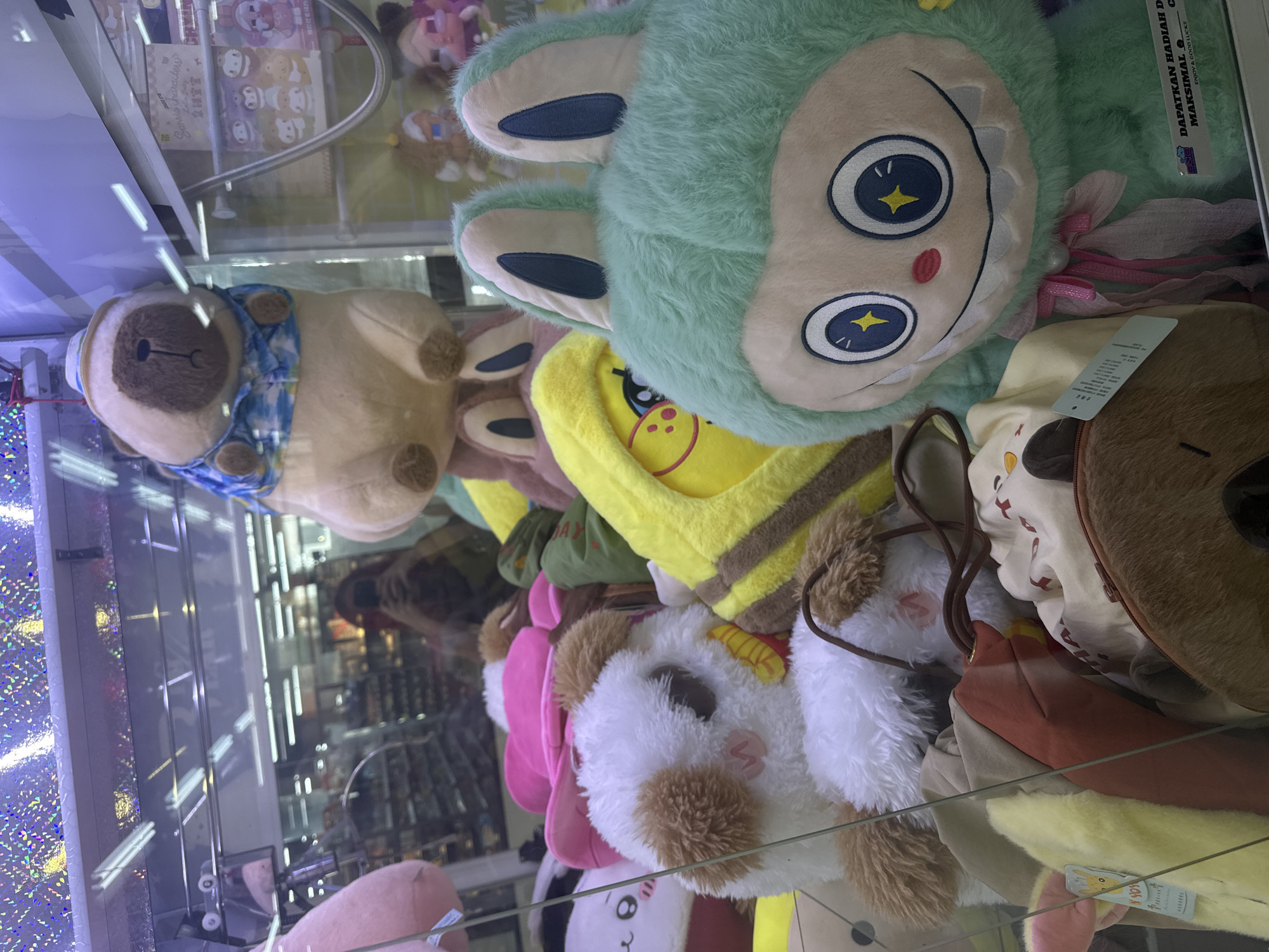 The image shows a claw machine filled with plush toys. In the foreground, a large, mint-green plush toy with a wide smile and large blue eyes with yellow stars is prominent. To its left is a white and brown plush dog with a pink bow. Behind these, a yellow plush toy resembling a banana character is visible. Further back, a tan pug plush toy wearing a blue and white patterned shirt and a straw hat sits atop other toys. The background shows a blurred reflection of a store interior with shelves of products and fluorescent lighting. The scene is inside a glass-enclosed claw machine, likely in a mall or entertainment center in North Kuta, Indonesia, as suggested by the text on the green plush toy which reads "DAPATKAN HADIAH DENGAN MAKSIMAL" (Get Gifts with Maximum) and "ENJOY & GOOD LUCK!!". There is no indication of people actively operating the machine in this particular frame, though reflections suggest a person taking the photo.