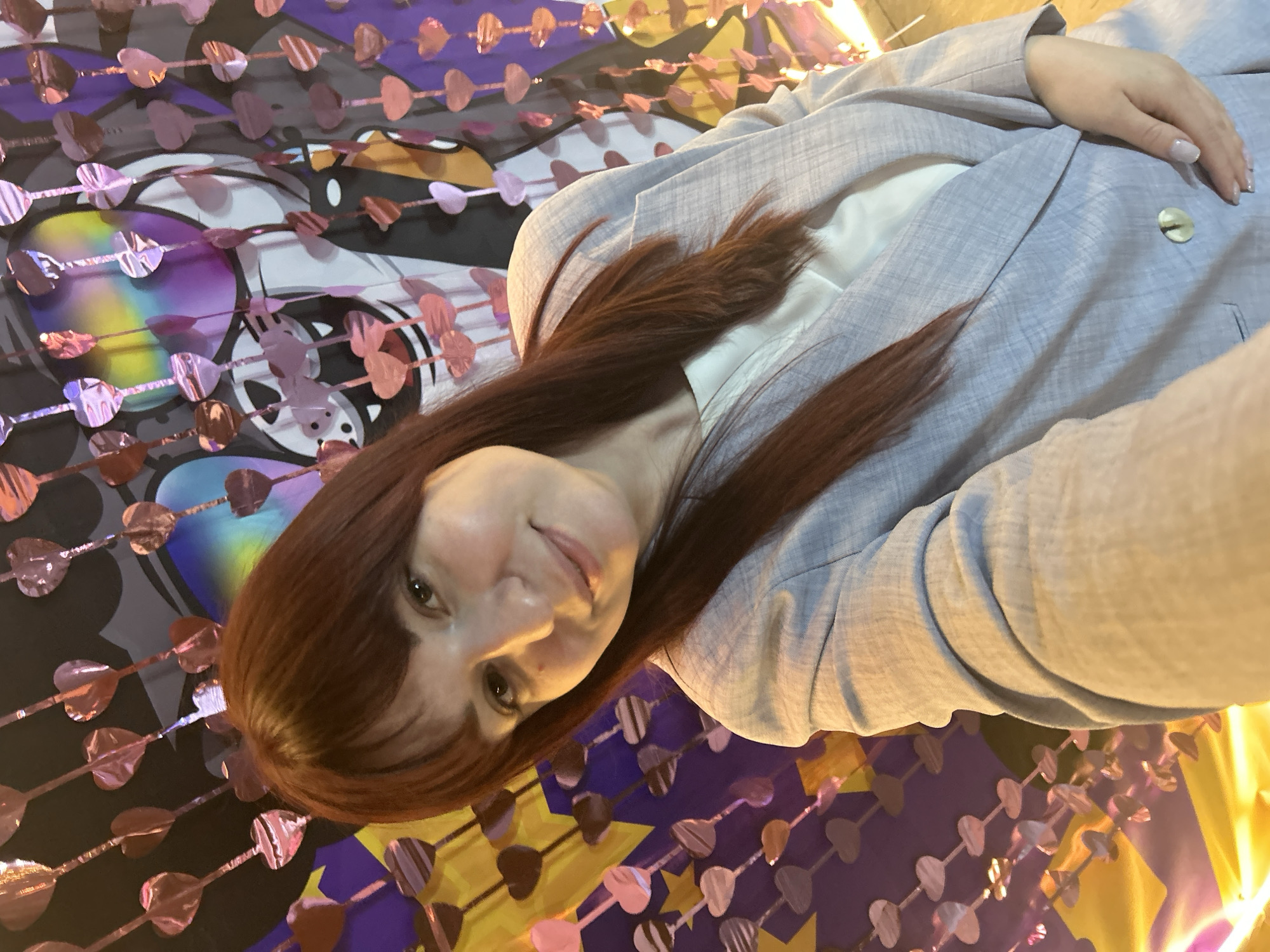 A woman with auburn hair is wearing a light blue blazer over a white shirt. She is positioned in front of a colorful backdrop featuring cartoonish designs and heart-shaped decorations. The picture appears to be taken in an indoor setting. Without more information, a specific location in Chaikovsky, Russia, is not determinable.
