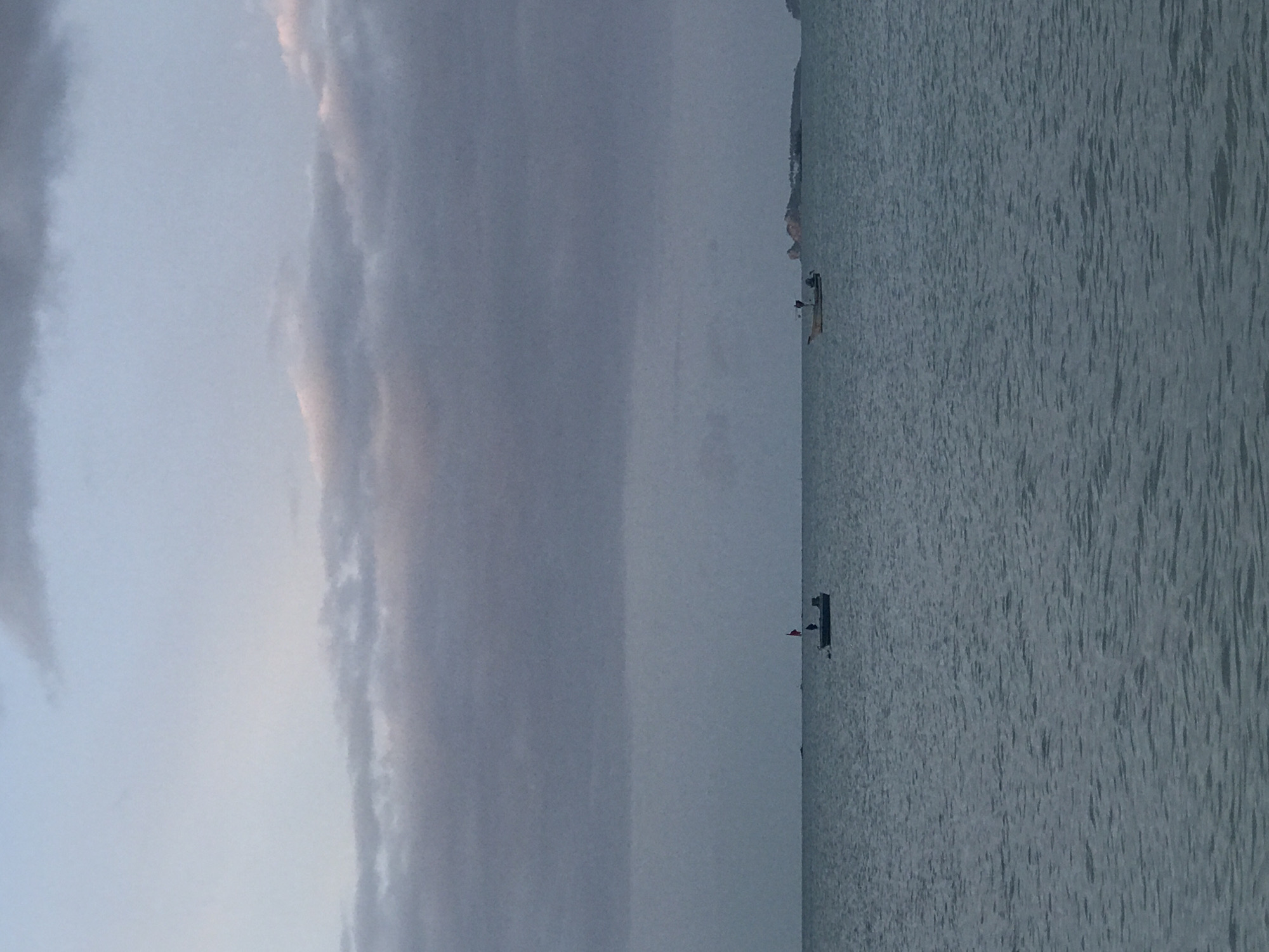 The image shows a body of water, likely the sea, under a cloudy sky. Two small boats are visible on the water. One boat, closer to the left side of the frame, appears to be dark and has a flag. The second boat, to the right, is lighter in color and also has a flag. In the distance, on the horizon, land or islands are visible. The water is choppy, indicating some wind. The sky is overcast with clouds, suggesting it might be dawn or dusk. The scene is peaceful and serene. The location is in Baan Mae Nam, Thailand.