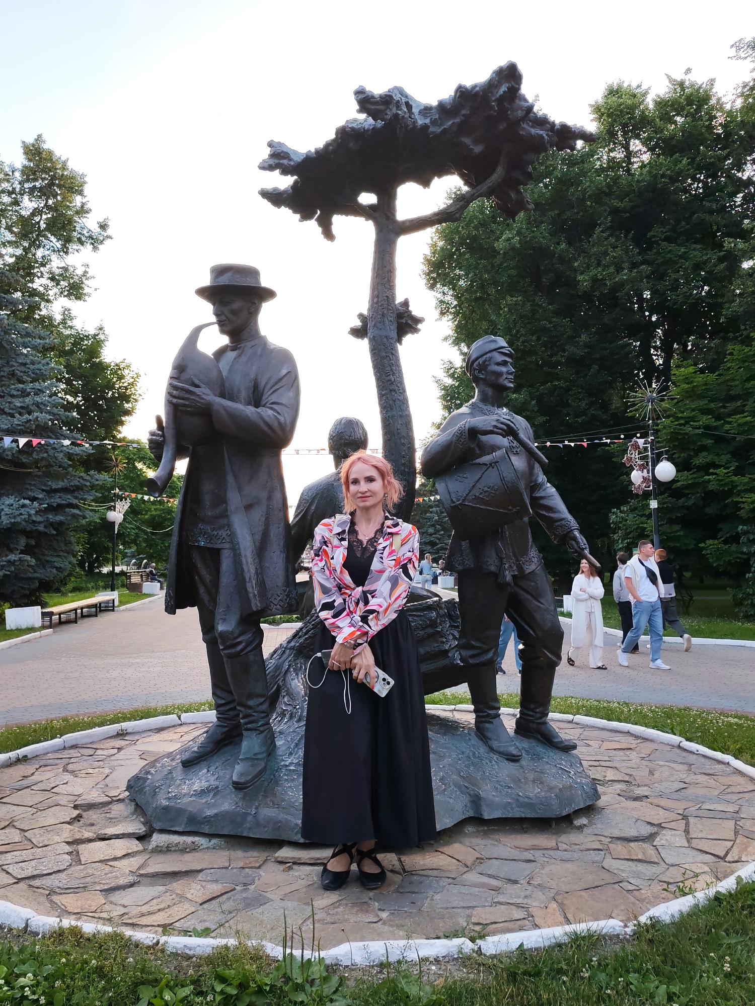 A woman stands in front of a bronze sculpture in Yoshkar-Ola, Russia. The sculpture depicts three figures, two men playing musical instruments, and a third figure partially obscured behind them. The man on the left wears a hat and plays a bagpipe-like instrument, while the man on the right plays a drum. A tree-like structure rises behind the figures. The woman, with reddish-pink hair, wears a black dress and a colorful patterned blouse. She holds a smartphone and earbuds in her hands. The sculpture is located on a circular stone base. In the background, trees line a park-like setting with pathways and benches. Several other people are visible walking in the distance.
