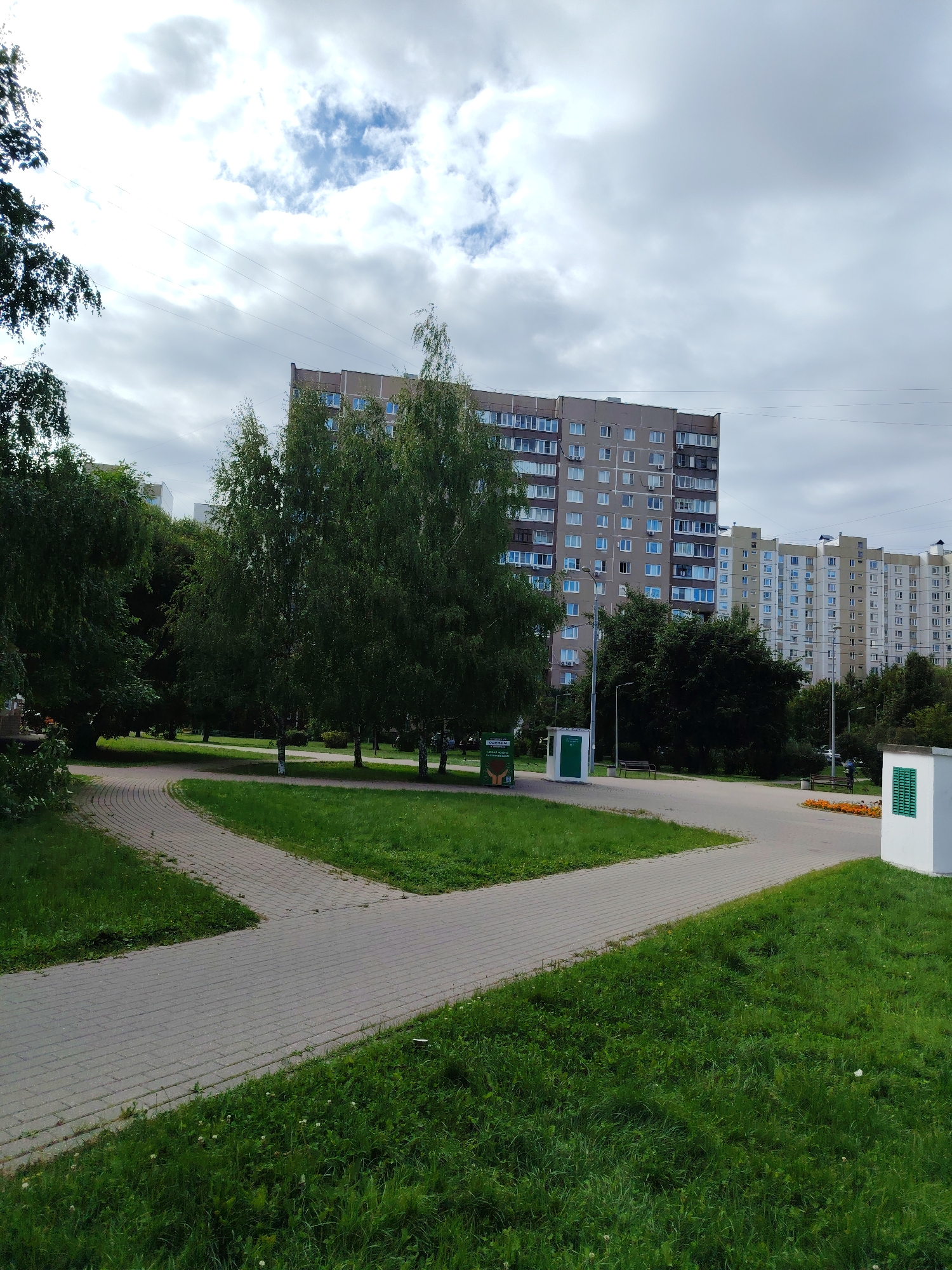 This image shows a view of a residential area in the city of Zelenograd, Russia. In the background, there are several multi-story apartment buildings, some of which appear to be older Soviet-era construction with beige and brown facades, while others are more modern with lighter colored facades. A winding paved walkway is visible in the foreground, bordered by well-maintained green grass lawns. Several deciduous trees with full green foliage are scattered throughout the scene, providing shade. A small green utility structure and a trash receptacle are positioned near the walkway. In the far right of the image, a white structure with green louvered windows is partially visible. The sky is overcast with a mix of clouds and some patches of blue. The overall impression is of a typical urban park or courtyard setting within a residential neighborhood.