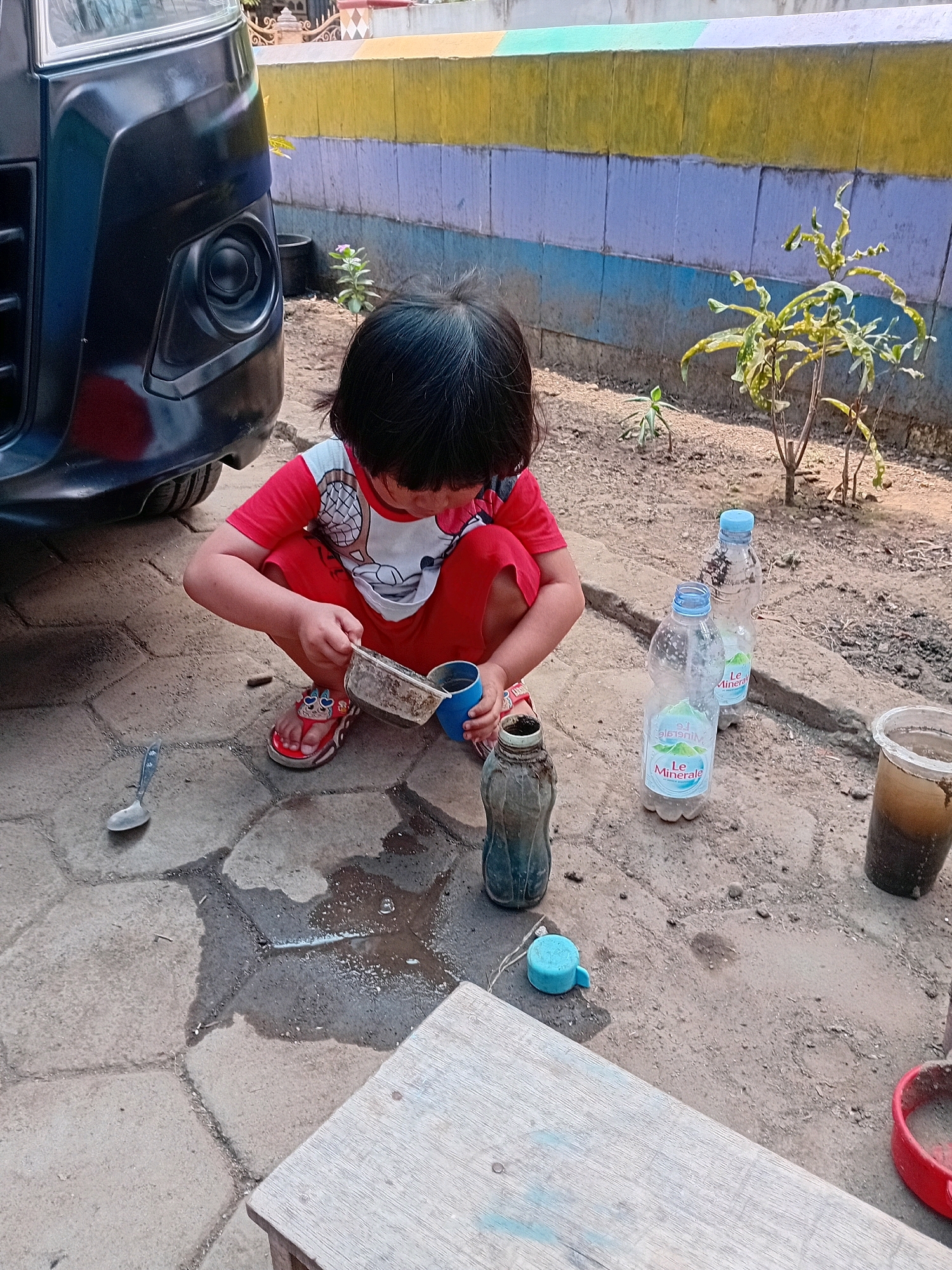 In Gebang, Indonesia, a young child with dark hair is crouched on a paved ground near a black vehicle. The child is wearing a red t-shirt with a graphic design and red shorts, along with red and white sandals. They are holding a grey container with a handle and pouring its contents into a blue cup. In front of the child, a metal spoon rests on the ground. To the right of the child, there are several plastic bottles, some of which are empty and others containing liquid. The background features a wall painted with horizontal stripes of yellow, light purple, and blue, and beyond that, dirt and sparse vegetation. The scene suggests a child playing outdoors with water and containers.