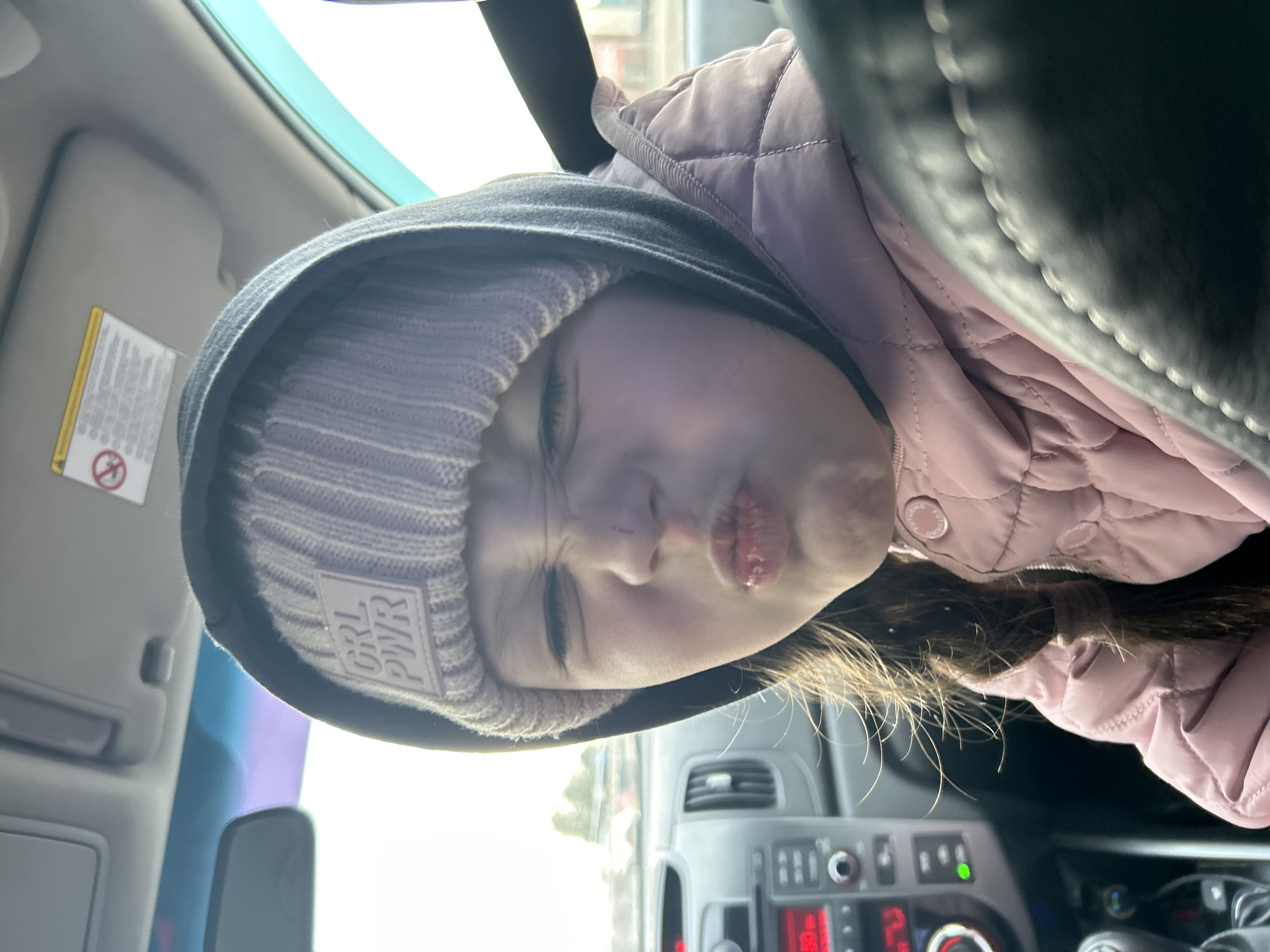 A young girl wearing a lavender beanie with "GRL PWR" on it and a black hoodie inside a car makes a kissing face at the camera. She is also wearing a pink quilted jacket. The car's dashboard is visible. The location cannot be determined with certainty as Chaikovsky, Russia with the provided information.