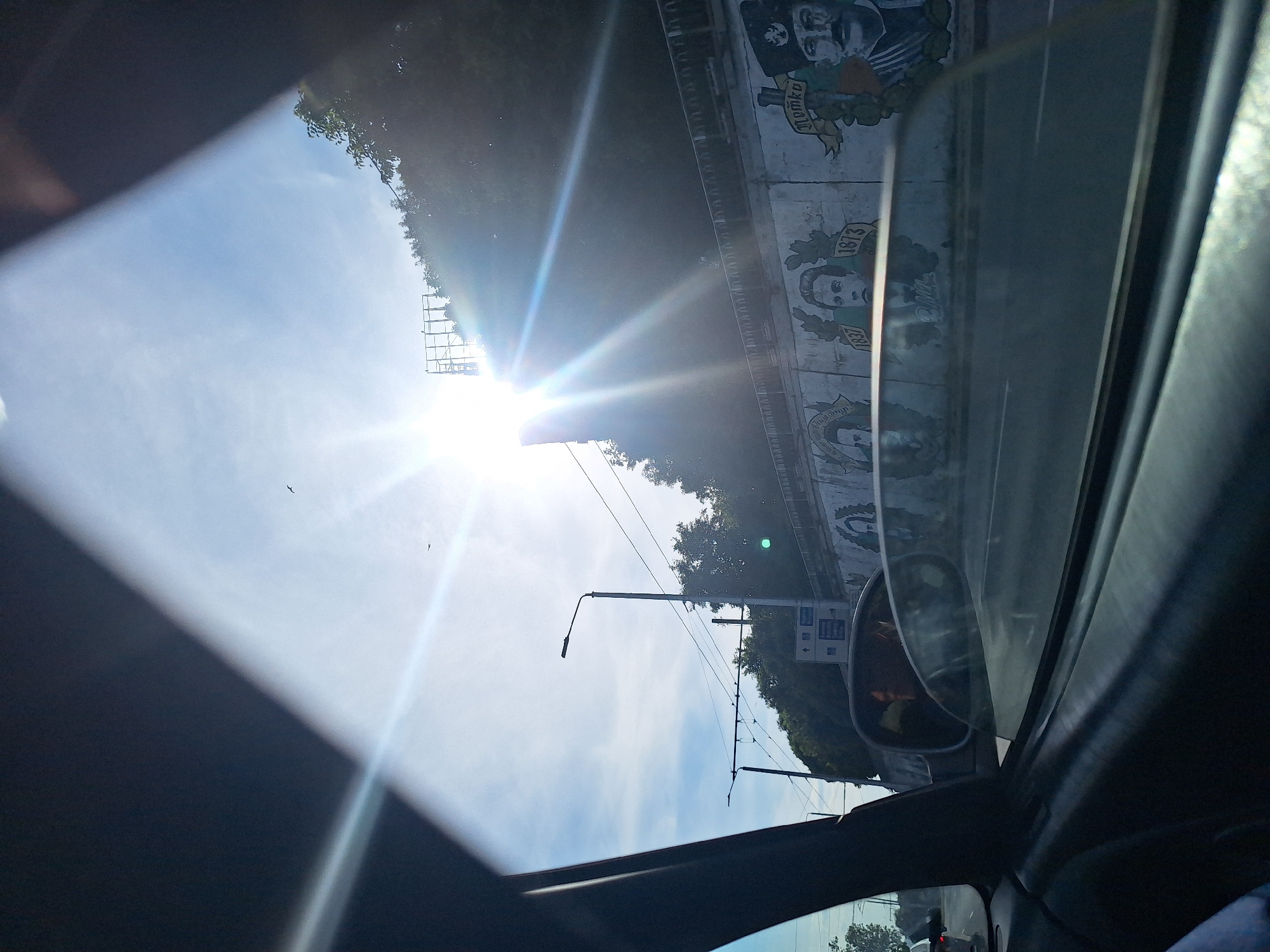 The image, taken from inside a vehicle looking out through the passenger side window and windshield, depicts a sunny day in Varna, Bulgaria. The sun is prominently displayed, casting strong rays and lens flare across the sky. The view encompasses a road with passing vehicles, a concrete wall adorned with murals, trees, and a bright sky with some clouds.
The murals on the wall appear to be portraits of historical figures, some accompanied by dates such as "1837" and "1873". The style suggests a patriotic or historical theme. A street lamp is visible on the left side of the frame. The side mirror of the car reflects a portion of the interior and possibly other vehicles or people. The overall environment is urban, with elements of infrastructure like the lamp posts and overhead wires indicating a functioning city. The sunlight suggests it is daytime, likely mid-morning or afternoon.
