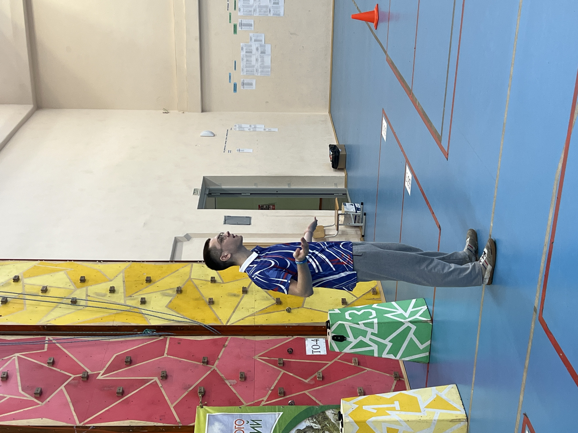 The image depicts a man standing in what appears to be an indoor sports facility, potentially a gym or climbing center in Berezniki, Russia. The walls are painted with climbing holds arranged on a red and yellow surface. A green geometric pattern box is near the man, and the blue floor has red lines marked on it. The man is wearing glasses, a blue shirt with white designs, and grey pants. He is facing the wall with his hands gesturing. In the background, there are papers attached to the wall, a doorway, and an orange cone on the blue floor.