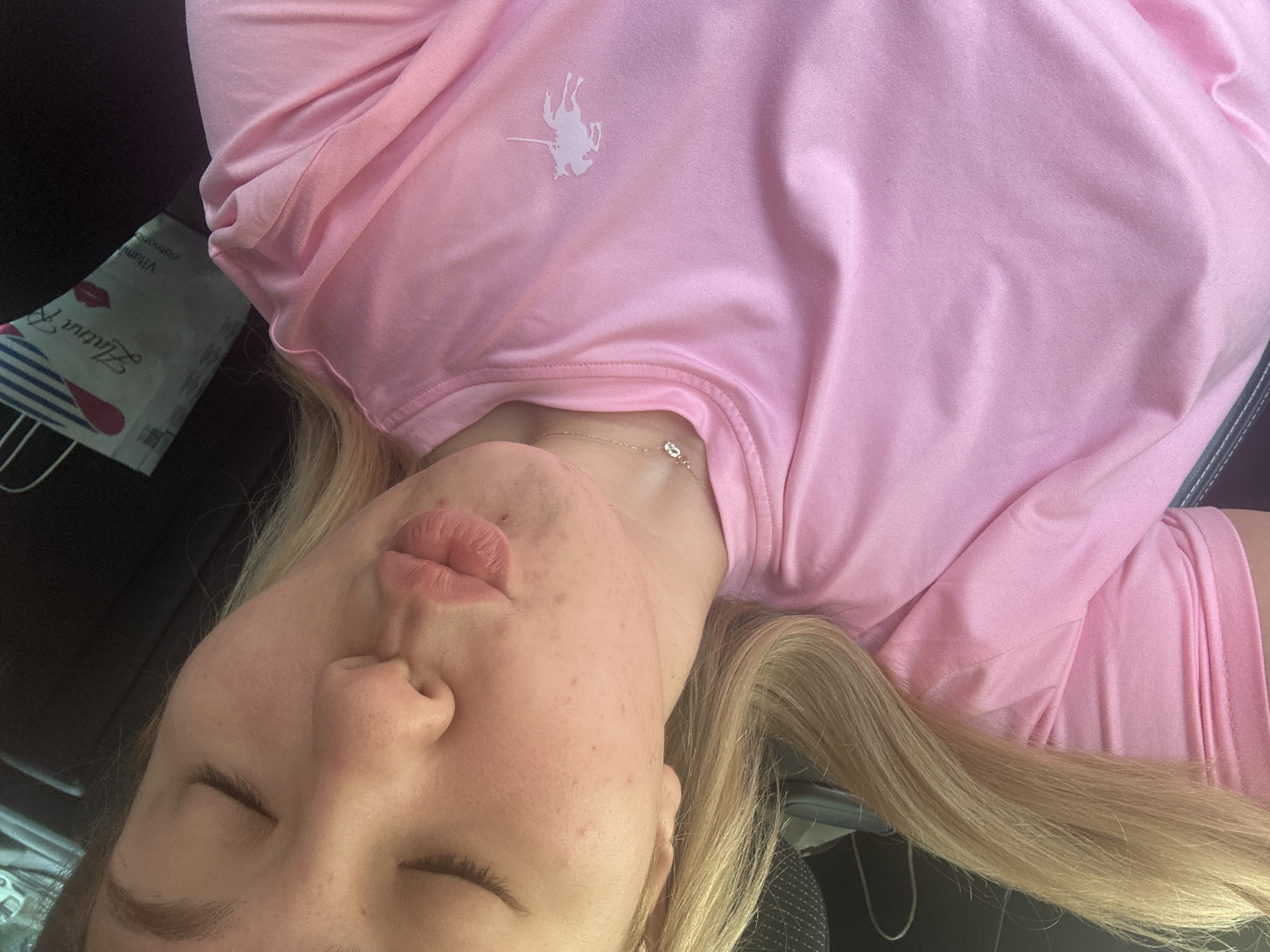 The image is a close-up, vertically oriented photograph of a young woman with blonde hair, her eyes closed, puckering her lips. She is wearing a pink t-shirt with a small white embroidered emblem of a polo player on a horse. A delicate silver necklace with a small pendant is visible around her neck. Her skin shows some minor blemishes. In the background, a portion of a bag with the text "Zlatna R" and an image of lips is visible. The context of Plovdiv, Bulgaria, is not evident from the image itself.