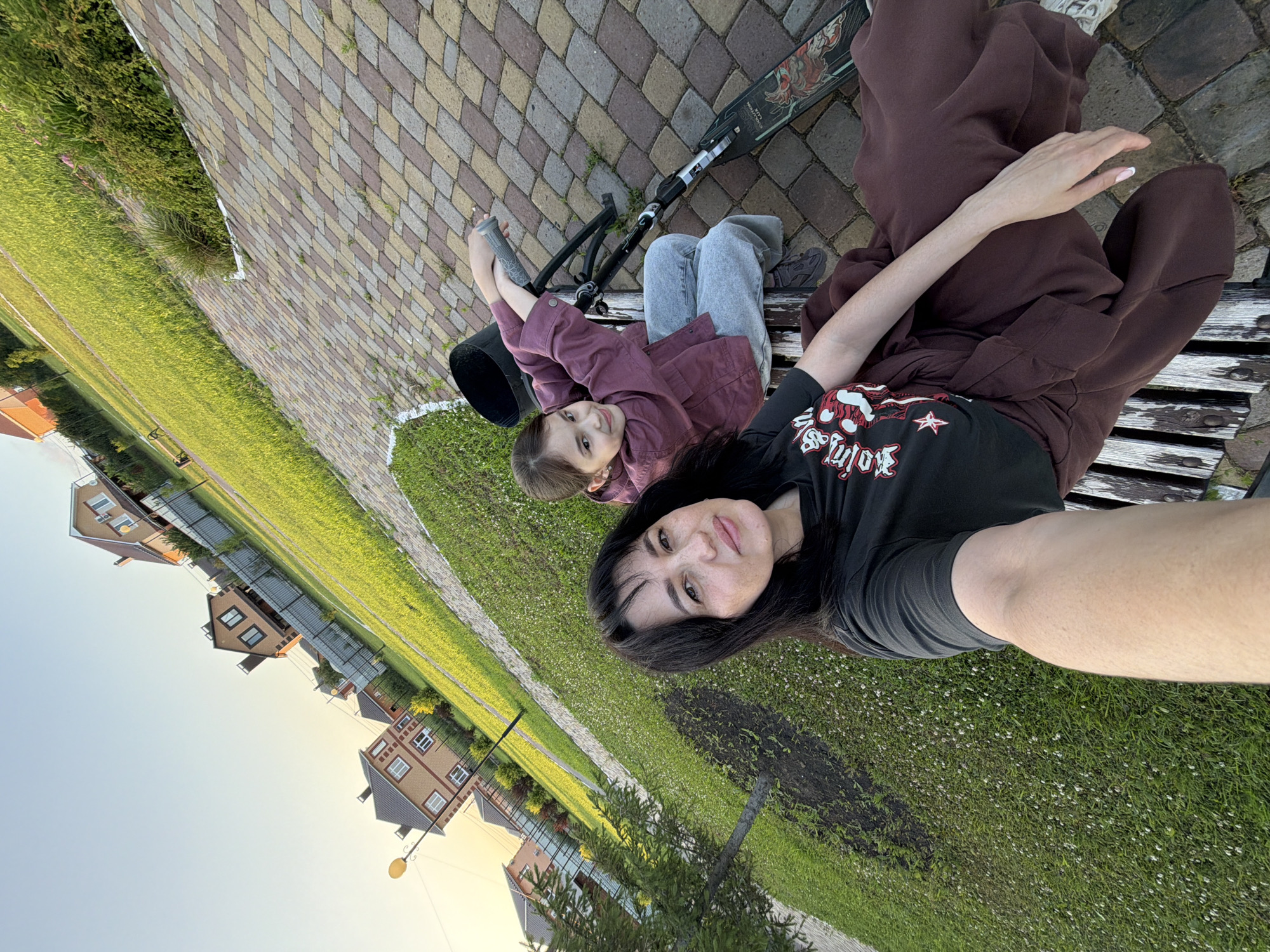 In Шмидтово, Russia, a woman and a young girl are sitting on a park bench. The woman, with dark hair and a black t-shirt, is in the foreground and is taking a selfie. The girl, wearing a maroon jacket and gray pants, is sitting next to her, smiling and resting her arms on the handlebars of a scooter parked beside them. The background shows a well-maintained lawn with scattered white flowers, a paved walkway, and several brick houses with fenced yards. The setting sun casts a warm glow on the scene.
