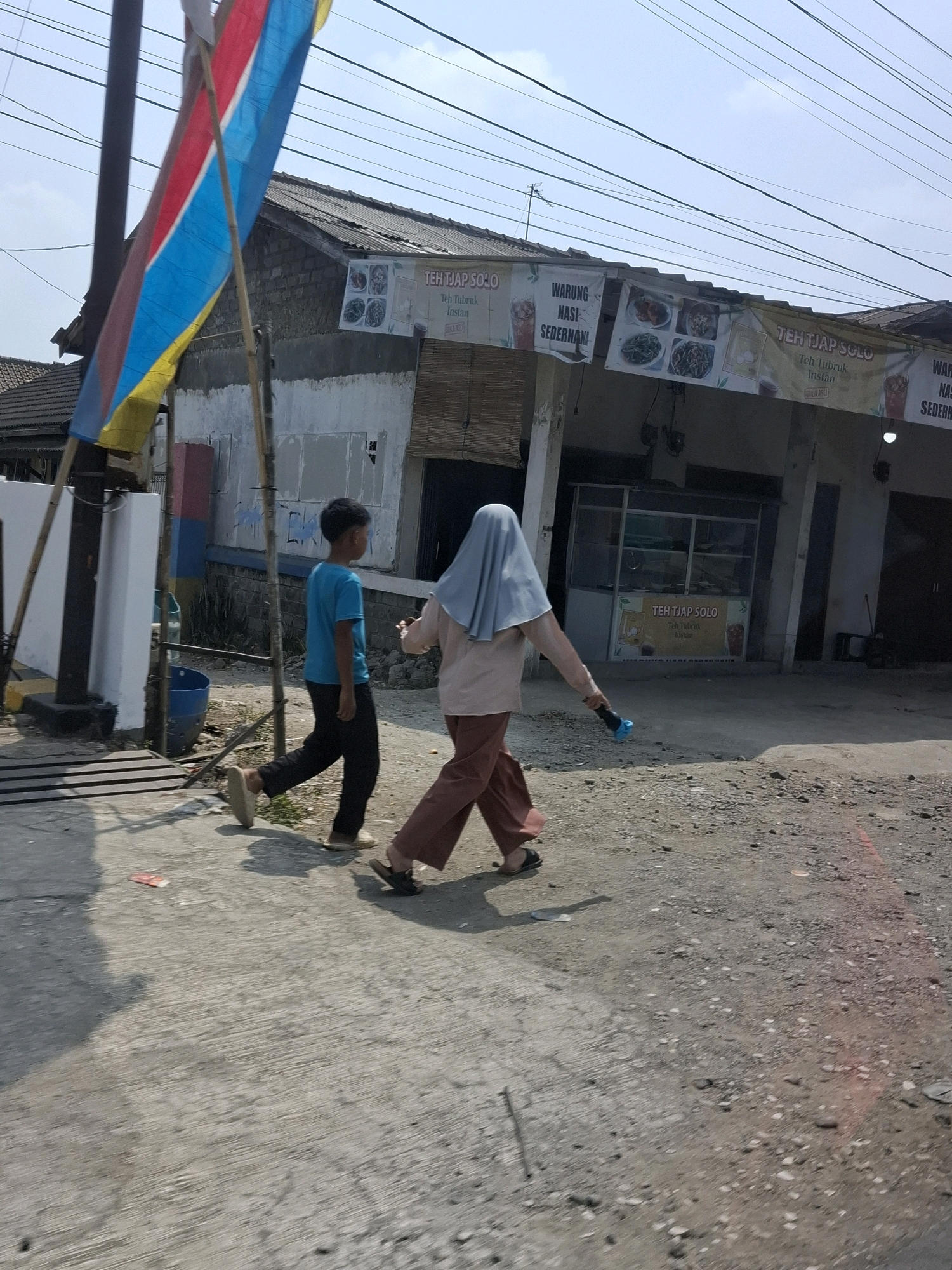 In Lembursawah, Indonesia, a street scene unfolds with two individuals walking along a paved and unpaved surface. The person on the left, a young boy with dark hair, wears a blue t-shirt and black trousers, and is barefooted. To his right, an individual, likely an adult, is dressed in a light pink long-sleeved shirt and loose brown trousers, with a light blue hijab covering their head. They are wearing sandals and carrying a blue object in their right hand. In the background, a building with a corrugated metal roof stands, adorned with several banners displaying text and images of food and drinks. A colorful flag, predominantly red, blue, and yellow, flutters on a pole to the left of the building. The environment appears to be an outdoor commercial area, with overhead power lines visible against a clear sky.