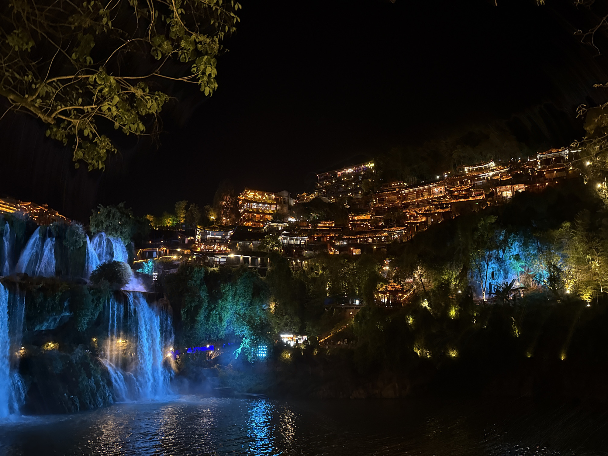 At night, a tiered waterfall flows powerfully into a dark river in Furong, China. The cascading water is illuminated with blue and white light. On the right, a hillside rises steeply, covered with traditional Chinese buildings, lit with warm yellow lights. Trees and vegetation surround the buildings, with some spots illuminated with blue, yellow, and white lights. In the foreground, the river reflects the lights from the waterfall and hillside. A tree branch with green leaves is visible in the upper left corner. The sky is dark.