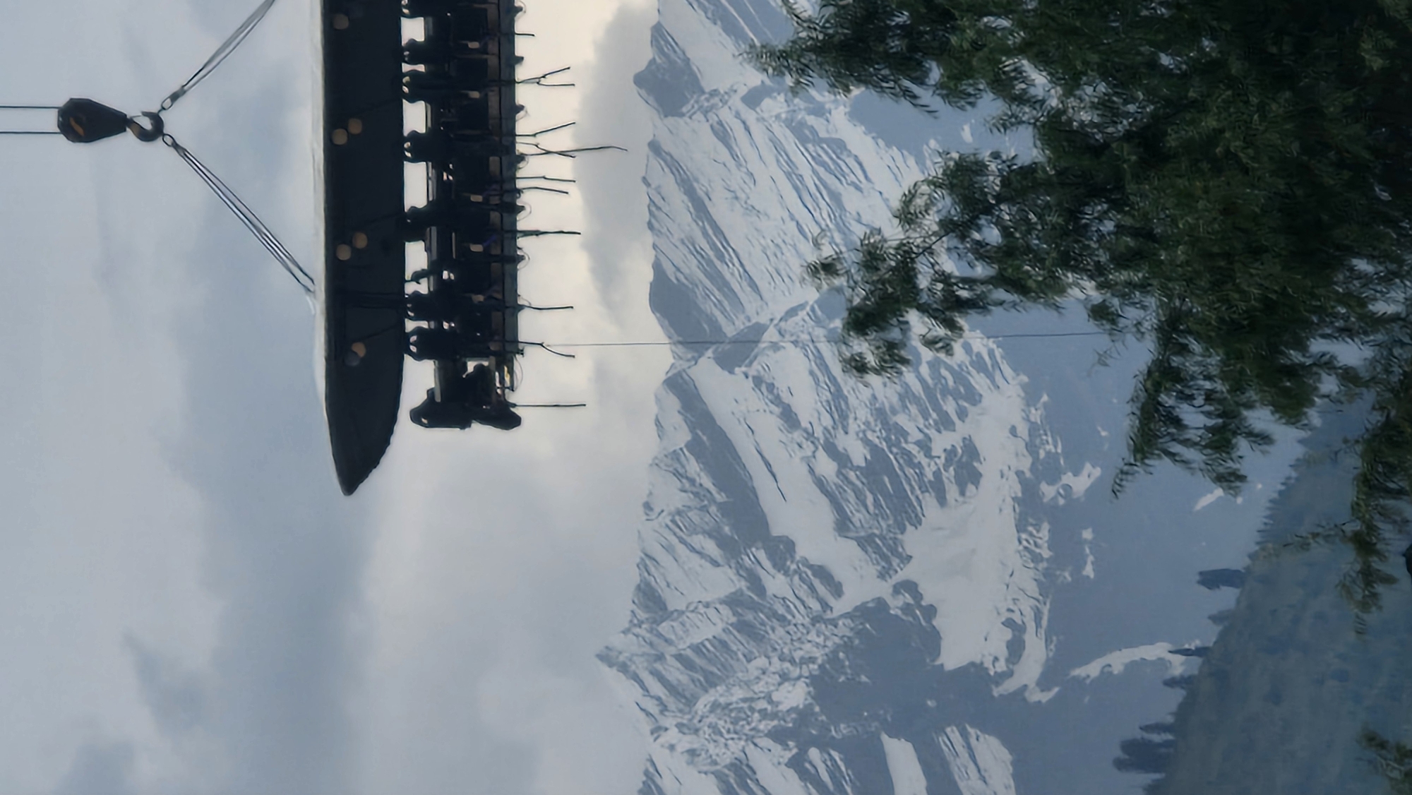 An aerial dining experience is suspended by a crane against a backdrop of snow-capped mountains in Vashisht, India. The dining platform, a large, dark, flat surface, is suspended at a significant height. Below the platform, chairs are arranged around a table, and numerous people are seated, appearing to be enjoying a meal or an experience. The platform is supported by thick cables attached to a crane hook visible at the top of the frame. In the background, majestic, rugged mountains dominate the landscape, their peaks covered in snow. The sky is overcast with grey clouds. In the foreground and mid-ground, lush green trees and hills are visible, indicating a natural, mountainous environment.
